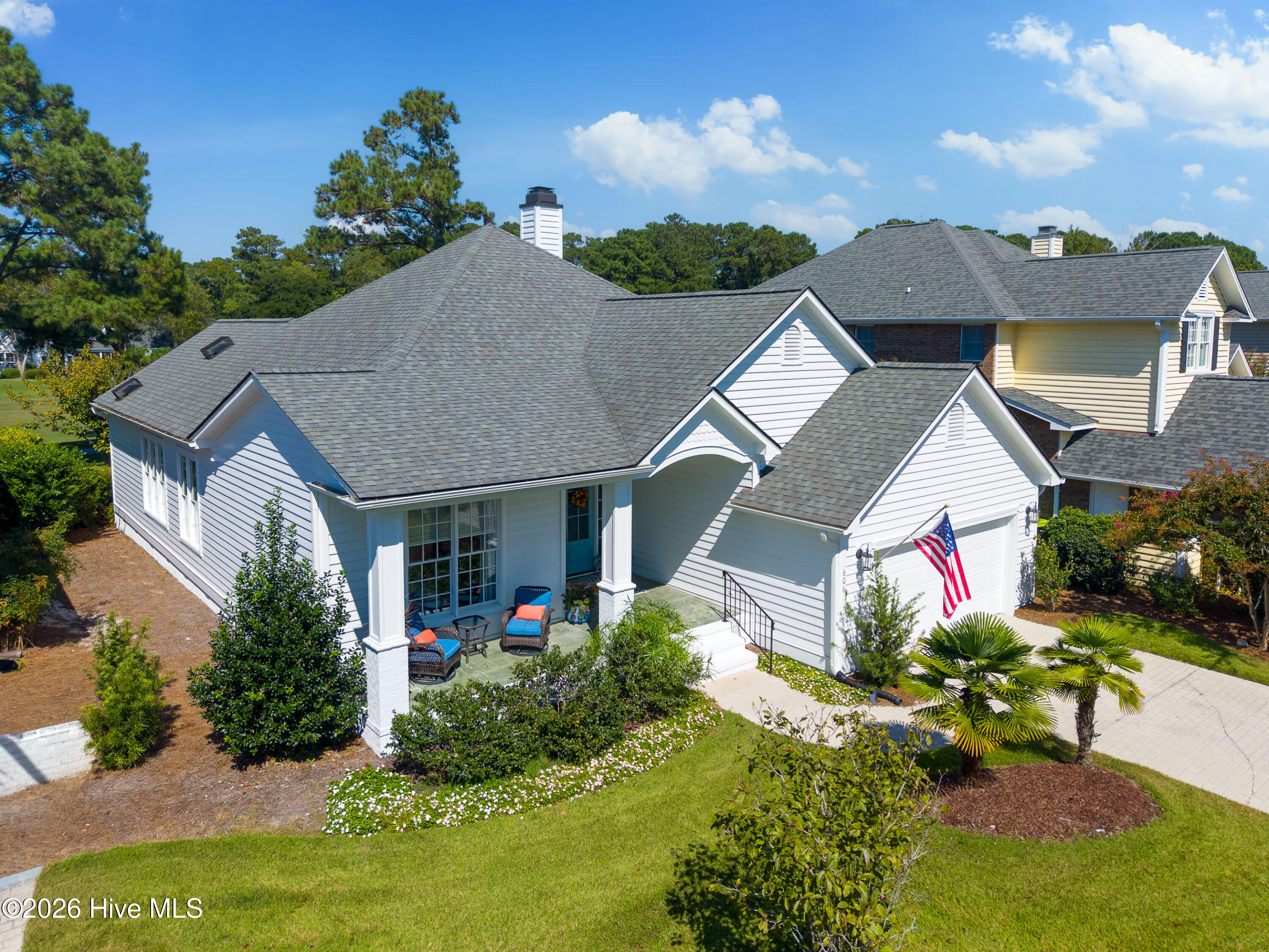 Light-gray house with a complex shingle roof, landscaped lawn, covered porch, and American flag beside the garage.