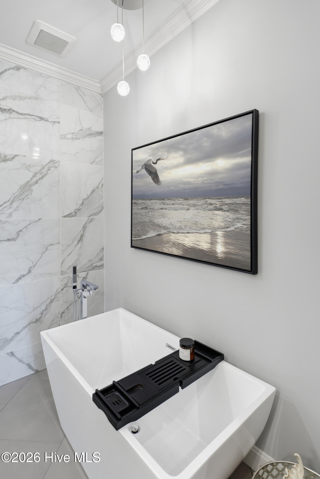 Freestanding white bathtub beside marble-look tile and framed coastal artwork.