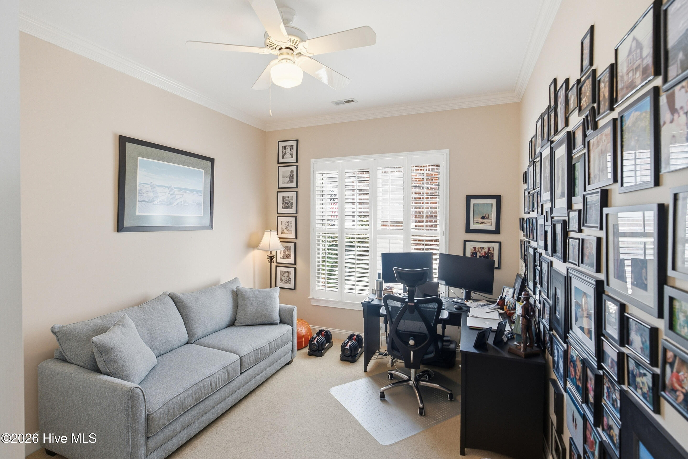Home office with a gray sofa, desk, plantation shutters, and a wall of framed photographs.