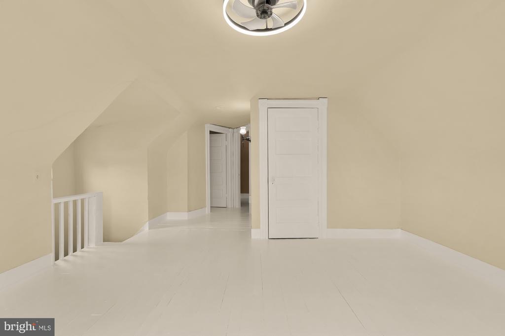 Vacant finished attic with sloped ceilings, white flooring, ceiling fan, and multiple doors.