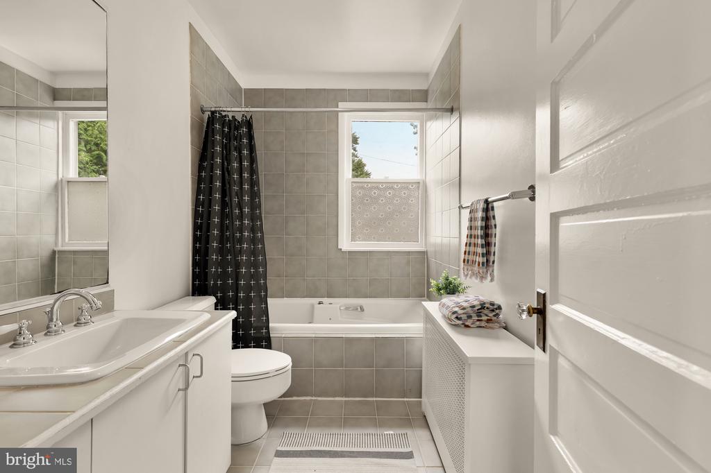 Bathroom with gray tile, white bathtub, vanity, and black patterned shower curtain.