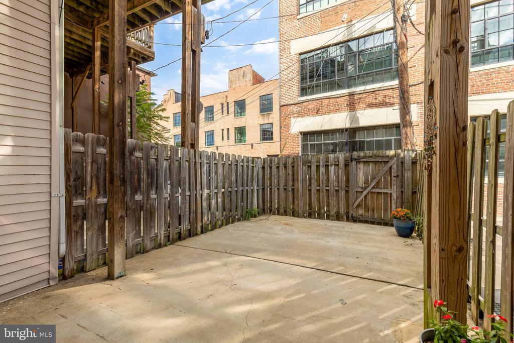 Fenced concrete patio beneath a raised wooden deck, with brick buildings beyond the fence.