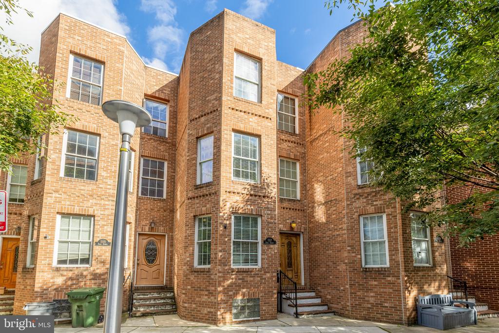 Red brick townhouse with tall windows, multiple front entrances, and leafy trees along the sidewalk.