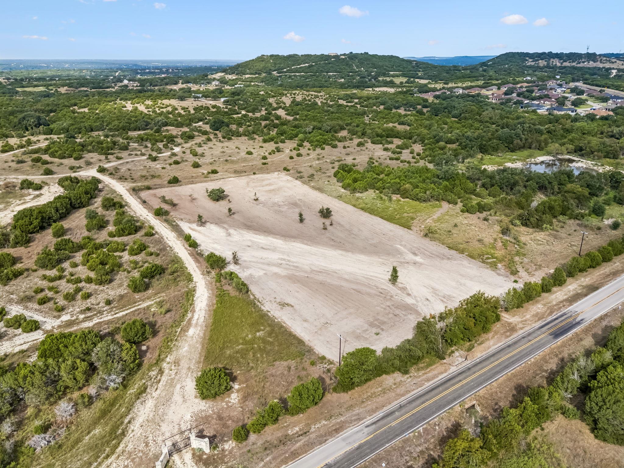 Aerial view of a cleared land parcel beside a two-lane road, with scrubland and hills beyond.