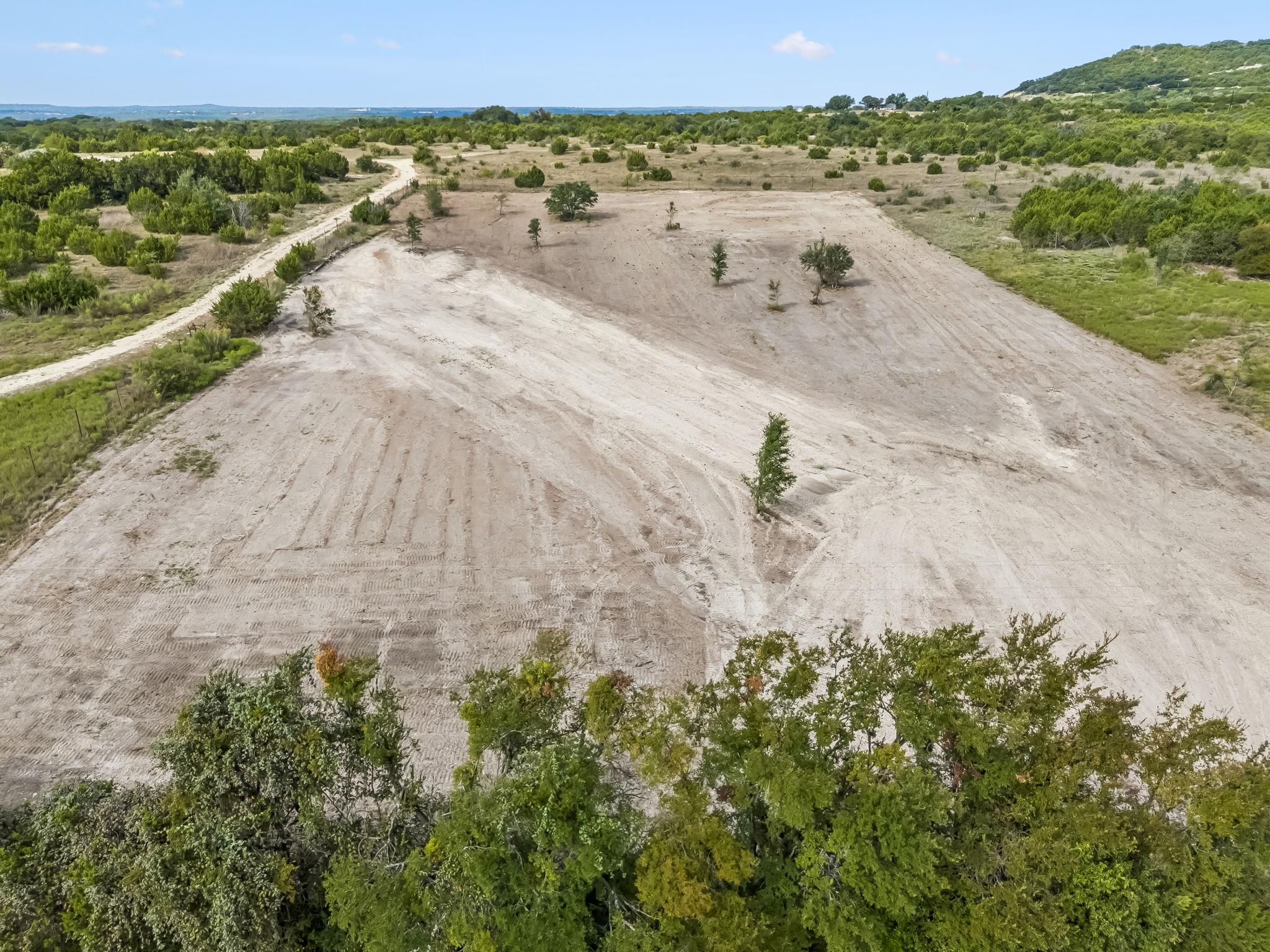 Aerial view of a cleared parcel with dirt tracks, scattered trees, and wooded hills beyond.