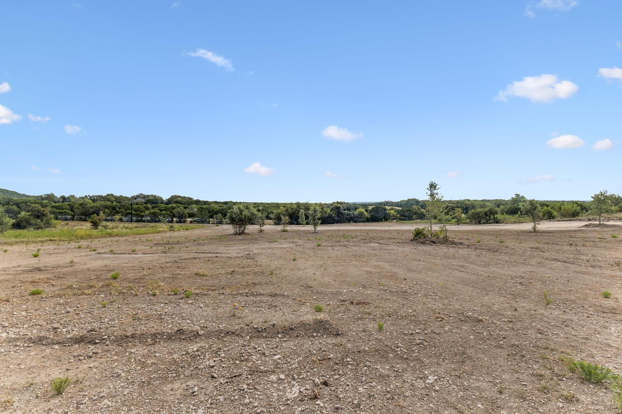 Cleared vacant land with rocky soil, scattered shrubs, and a distant tree line.