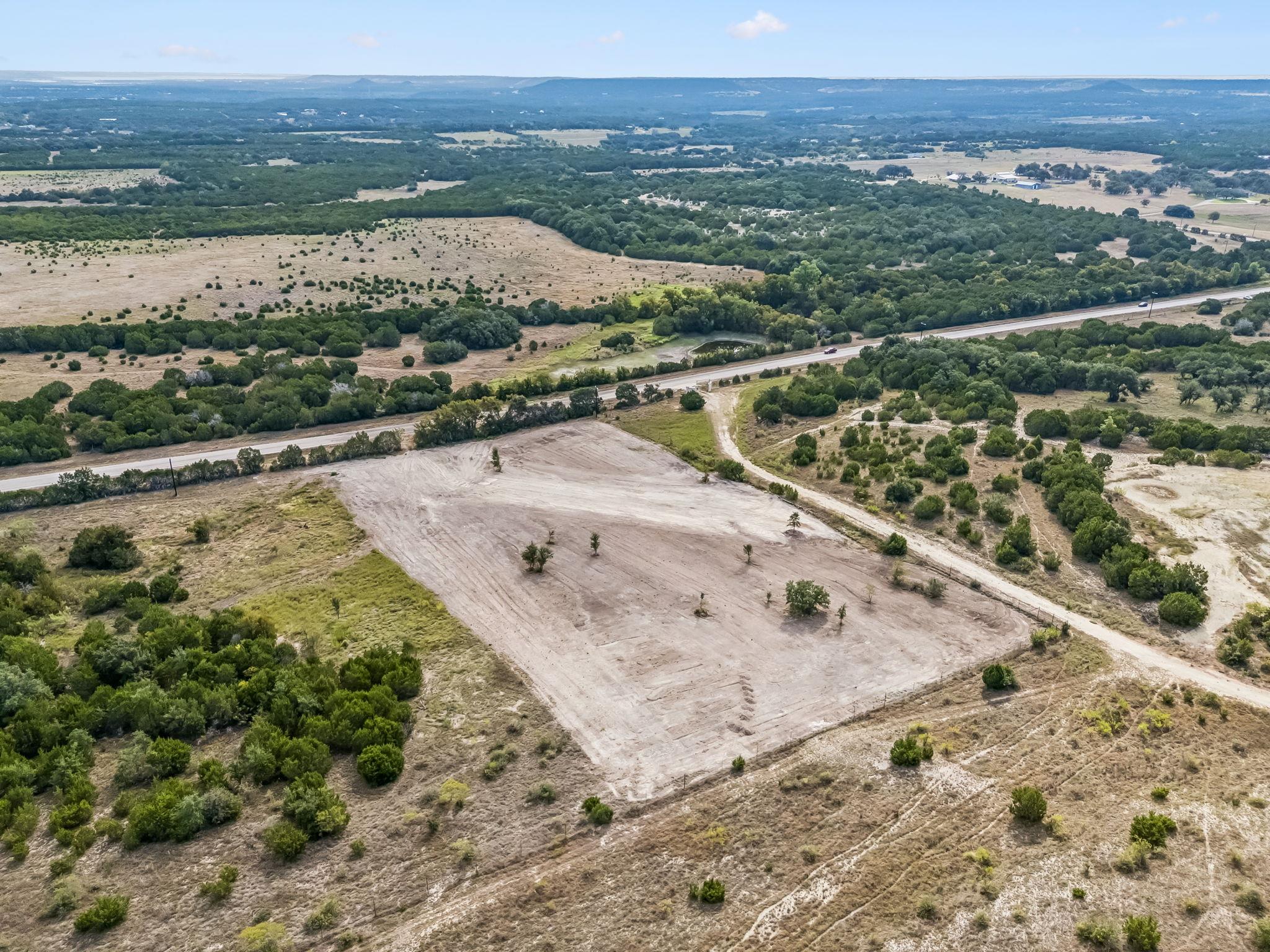 Cleared rural plot beside a paved road, surrounded by scrubland and trees.