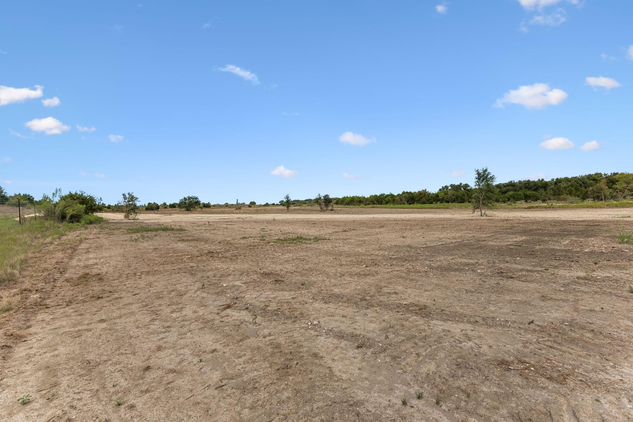 Cleared vacant land with dry soil, scattered trees, and a wooded horizon.