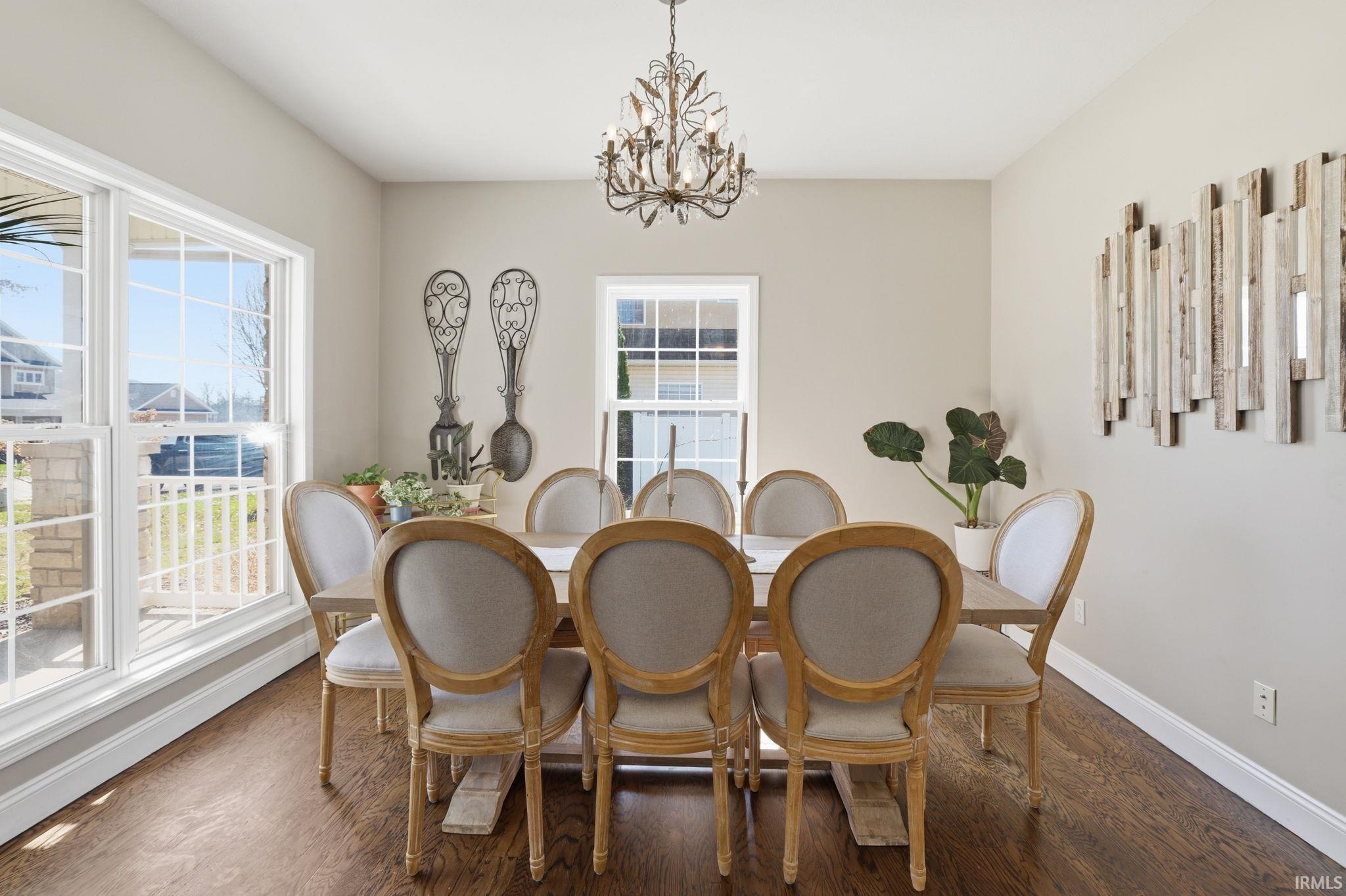 Dining room with a wooden table, upholstered chairs, chandelier, and tall windows.