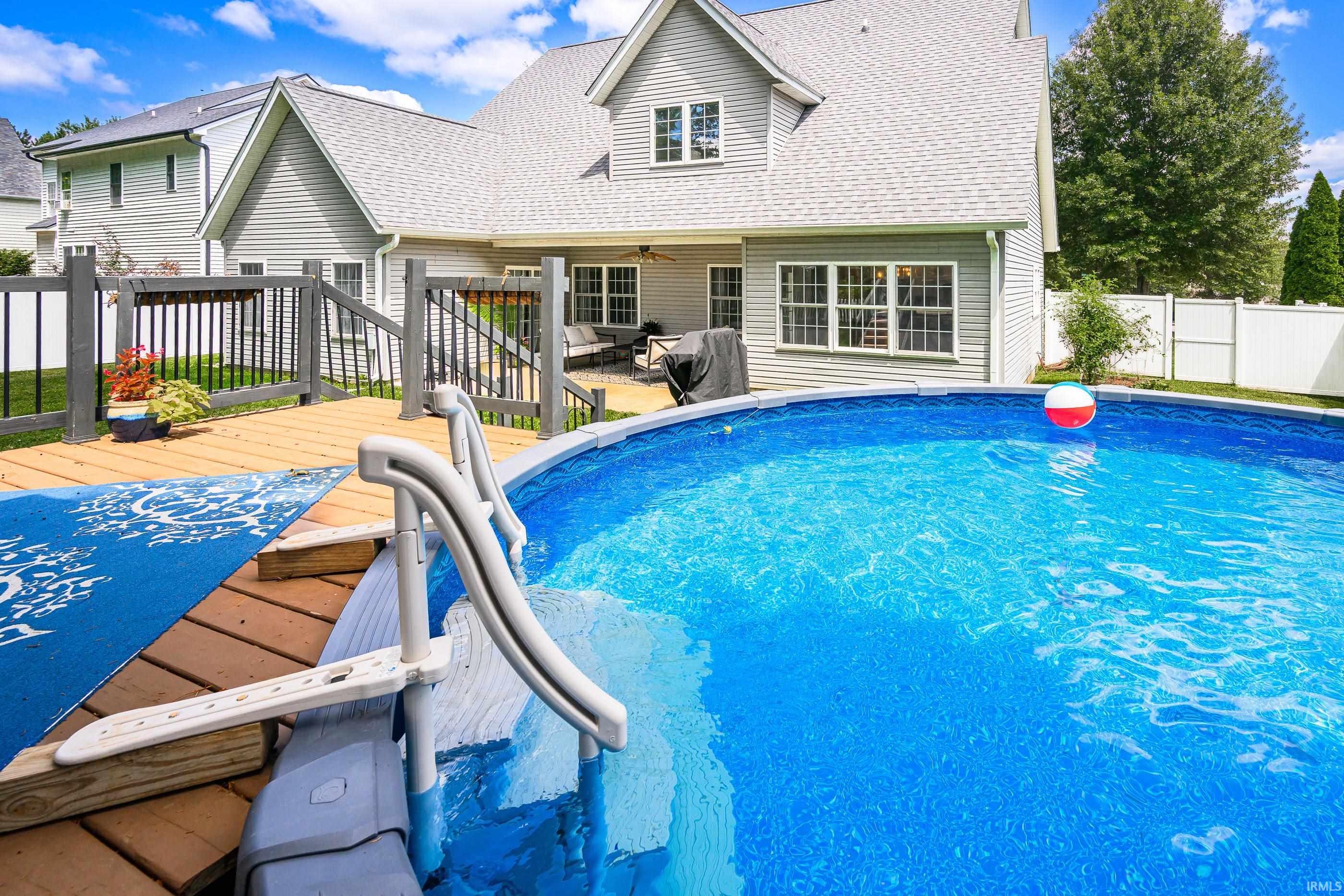 Backyard pool and wooden deck beside a two-story gray house, with patio seating and white fencing.