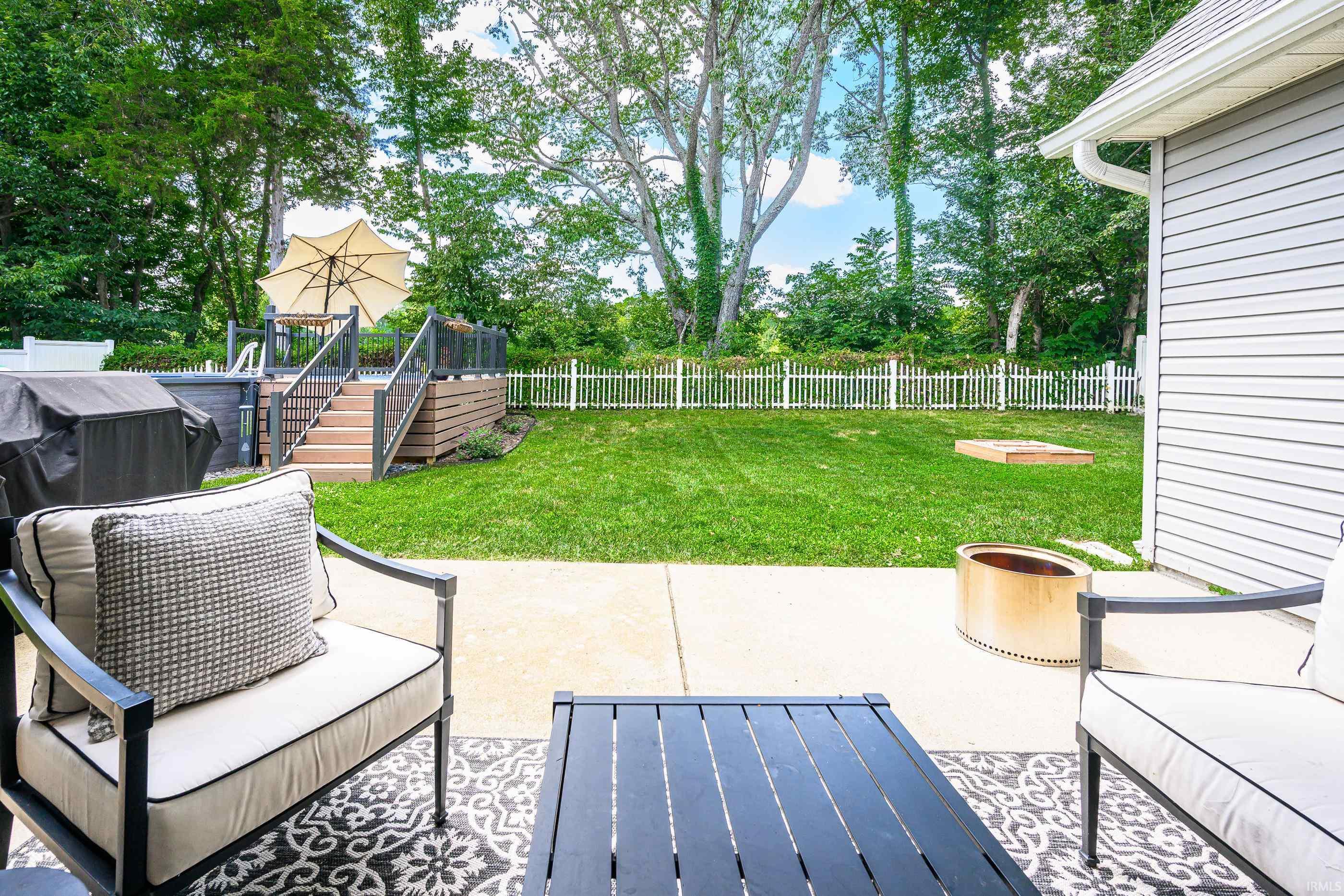 Backyard patio seating overlooks a fenced lawn, with a raised deck, umbrella, and fire pit.
