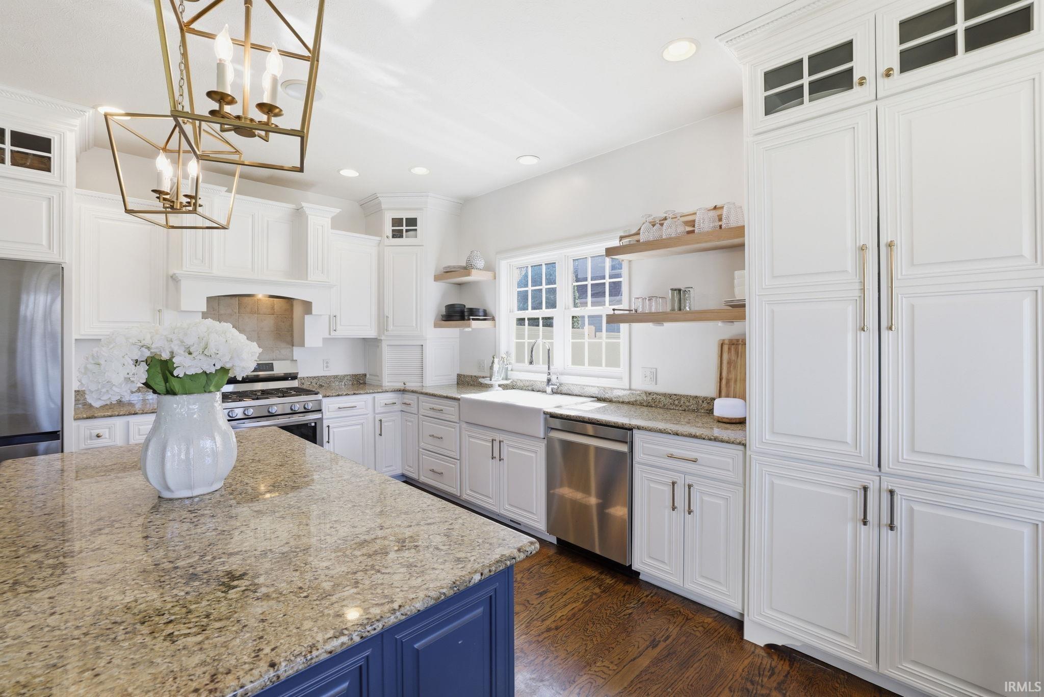 Bright kitchen with white cabinetry, granite counters, blue island, farmhouse sink, and brass pendant lights.