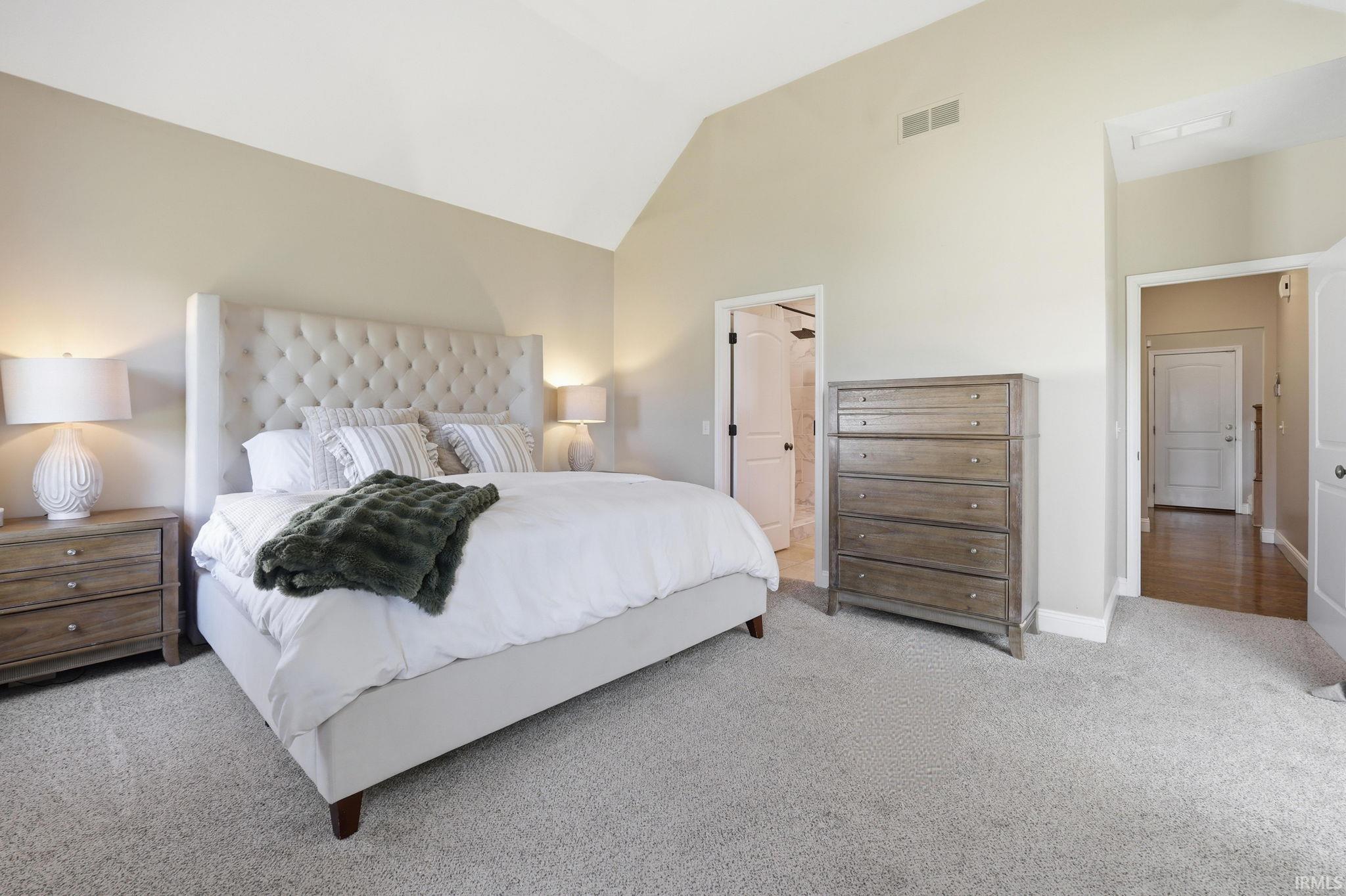 Staged bedroom with a white bed, tall tufted headboard, wood dressers, and vaulted ceiling.