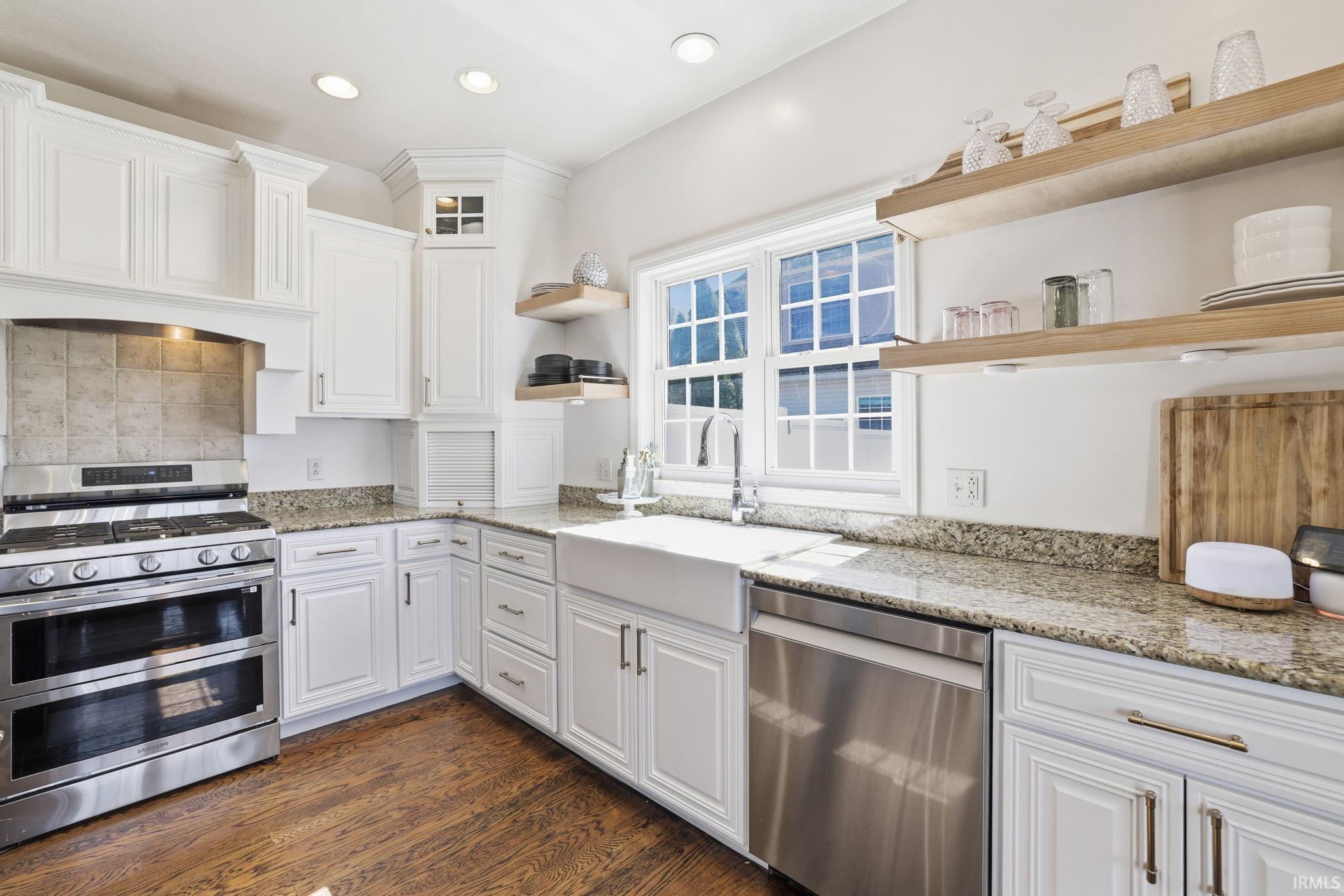 Bright white kitchen with granite counters, farmhouse sink, stainless steel appliances, and open wood shelves.