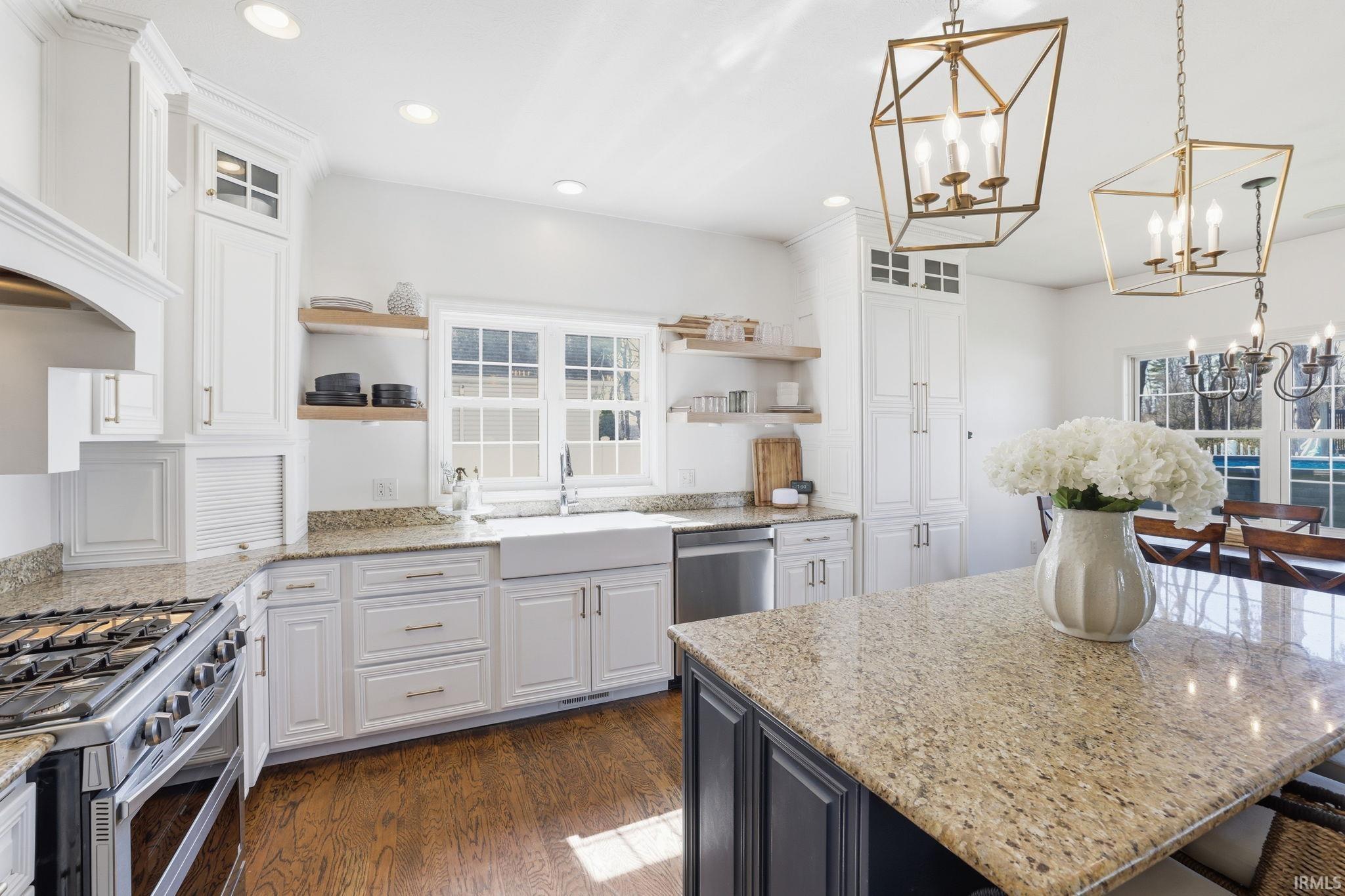Bright white kitchen with granite island, farmhouse sink, brass pendant lights, and a pool visible through the windows.