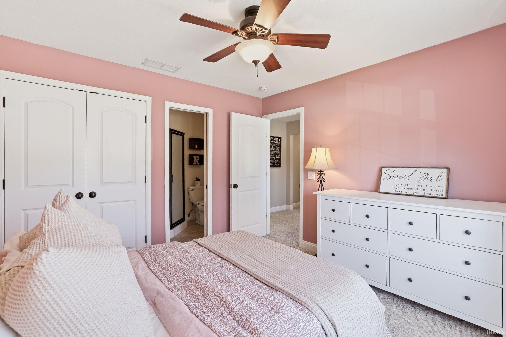 Bedroom with pink walls, patterned bedding, a white dresser, and a ceiling fan.