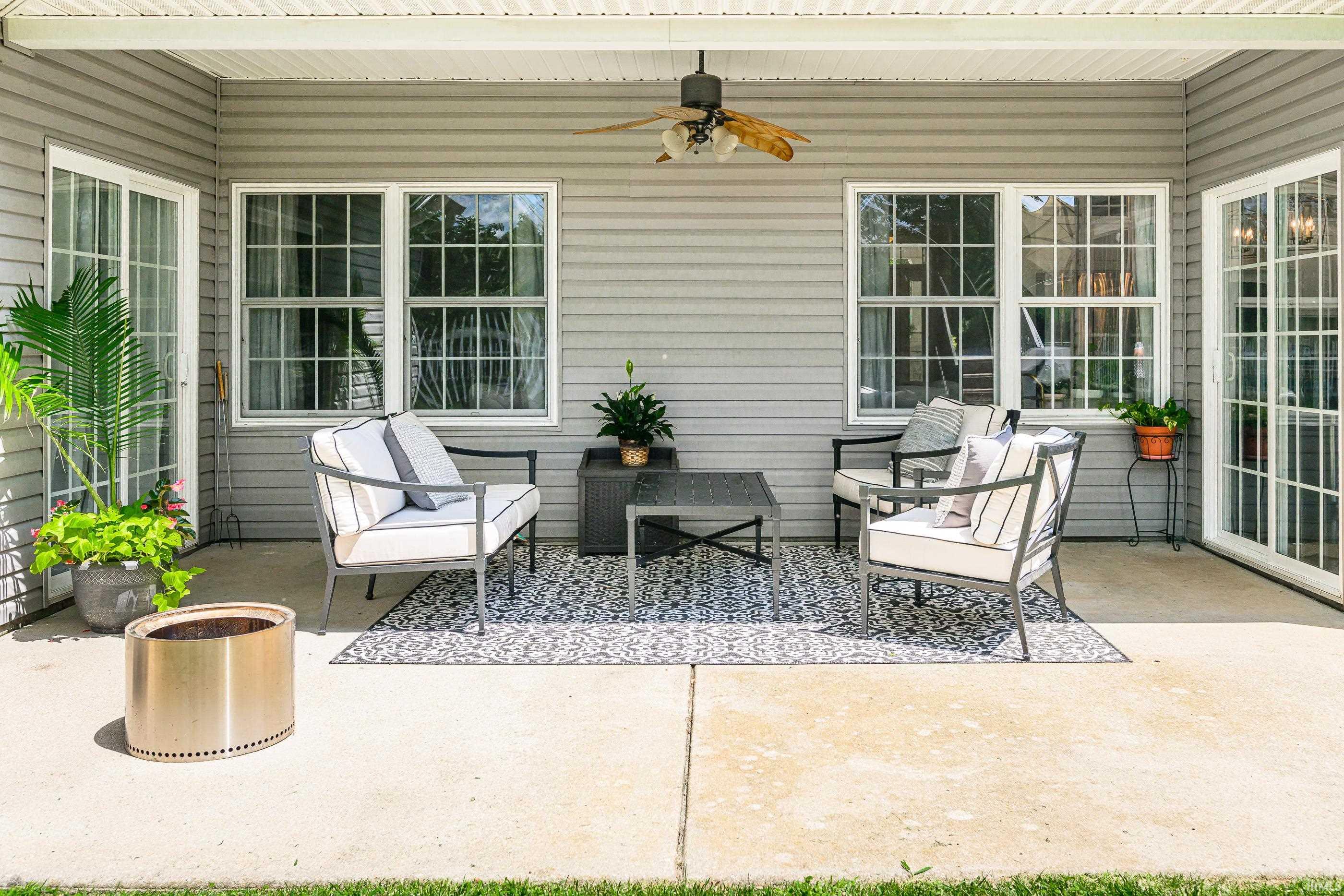 Covered patio with white-cushioned seating, patterned rug, ceiling fan, and potted plants.