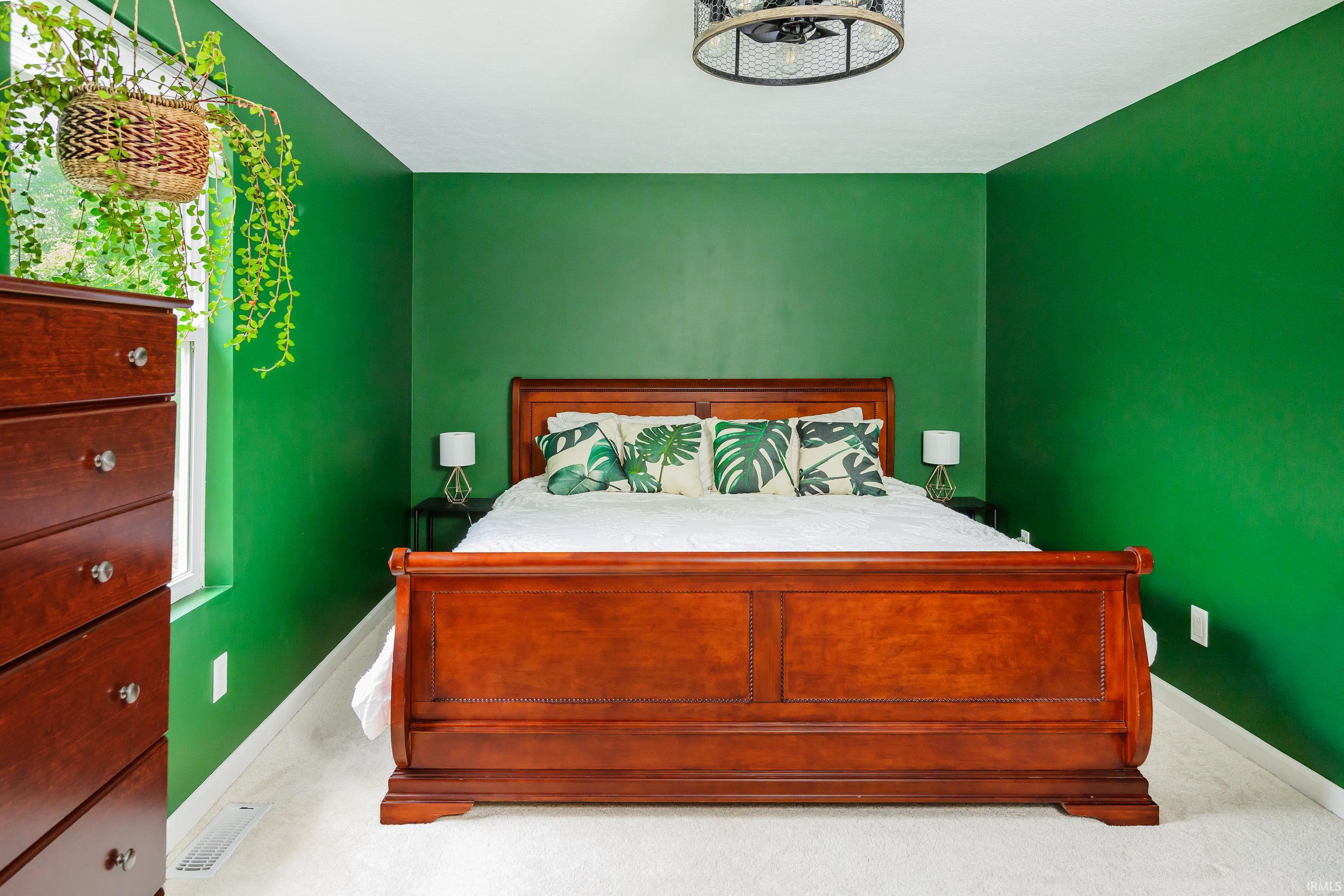 Bedroom with vivid green walls, a wooden bed, leaf-print pillows, and a hanging plant by the window.