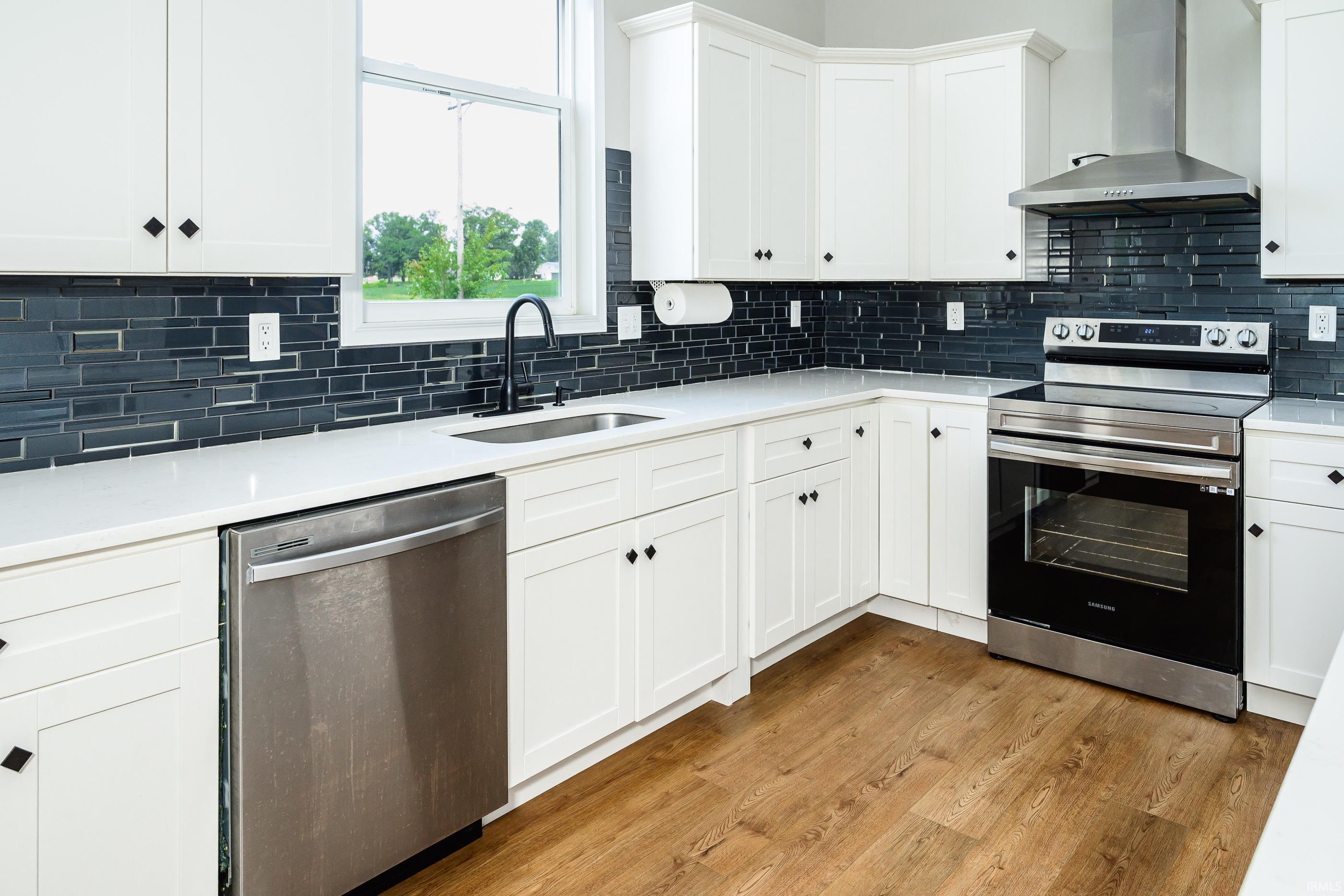 Renovated kitchen with white shaker cabinets, dark tile backsplash, stainless appliances, and warm wood flooring.