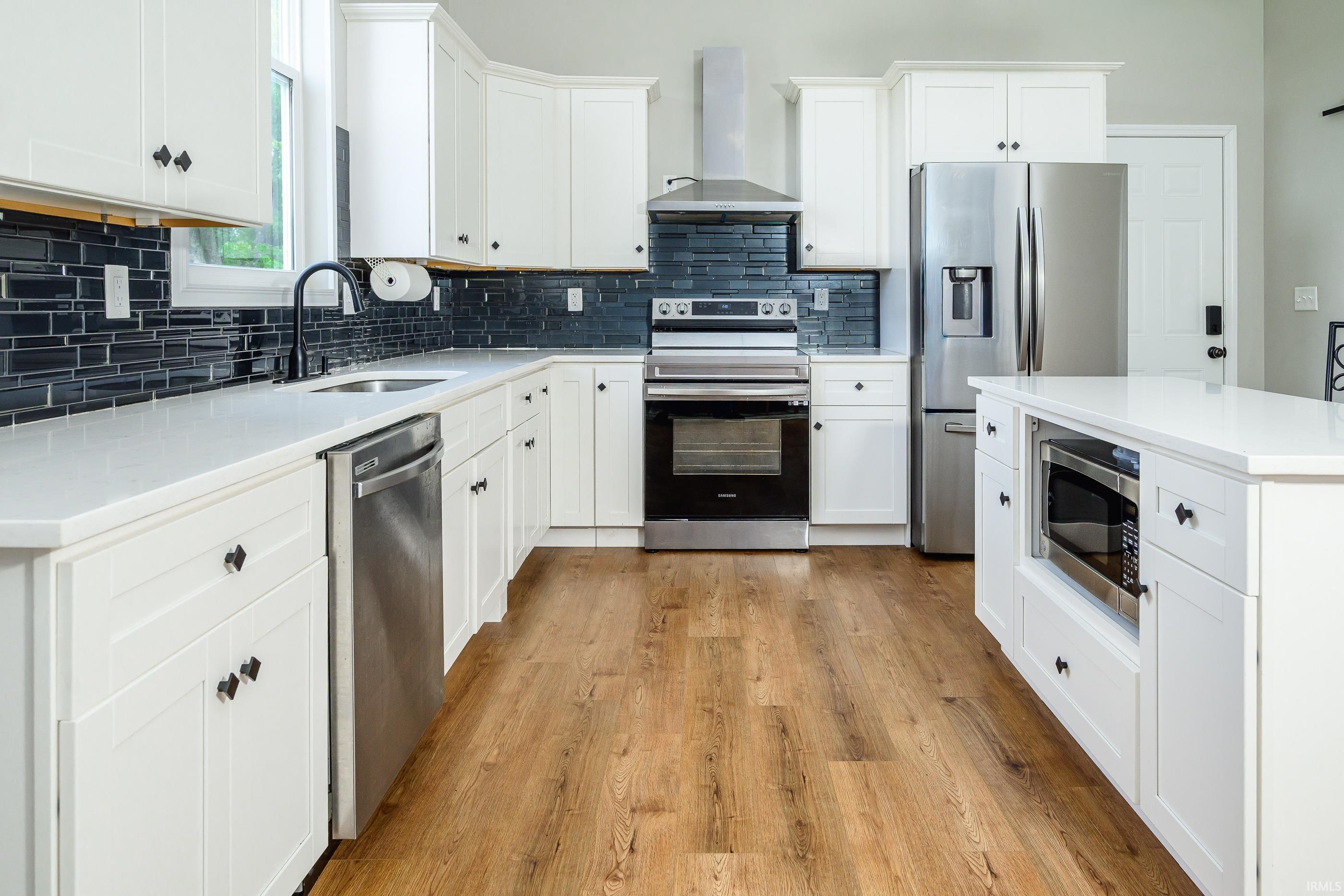 Kitchen with white shaker cabinets, black tile backsplash, stainless steel appliances, and warm wood floors.