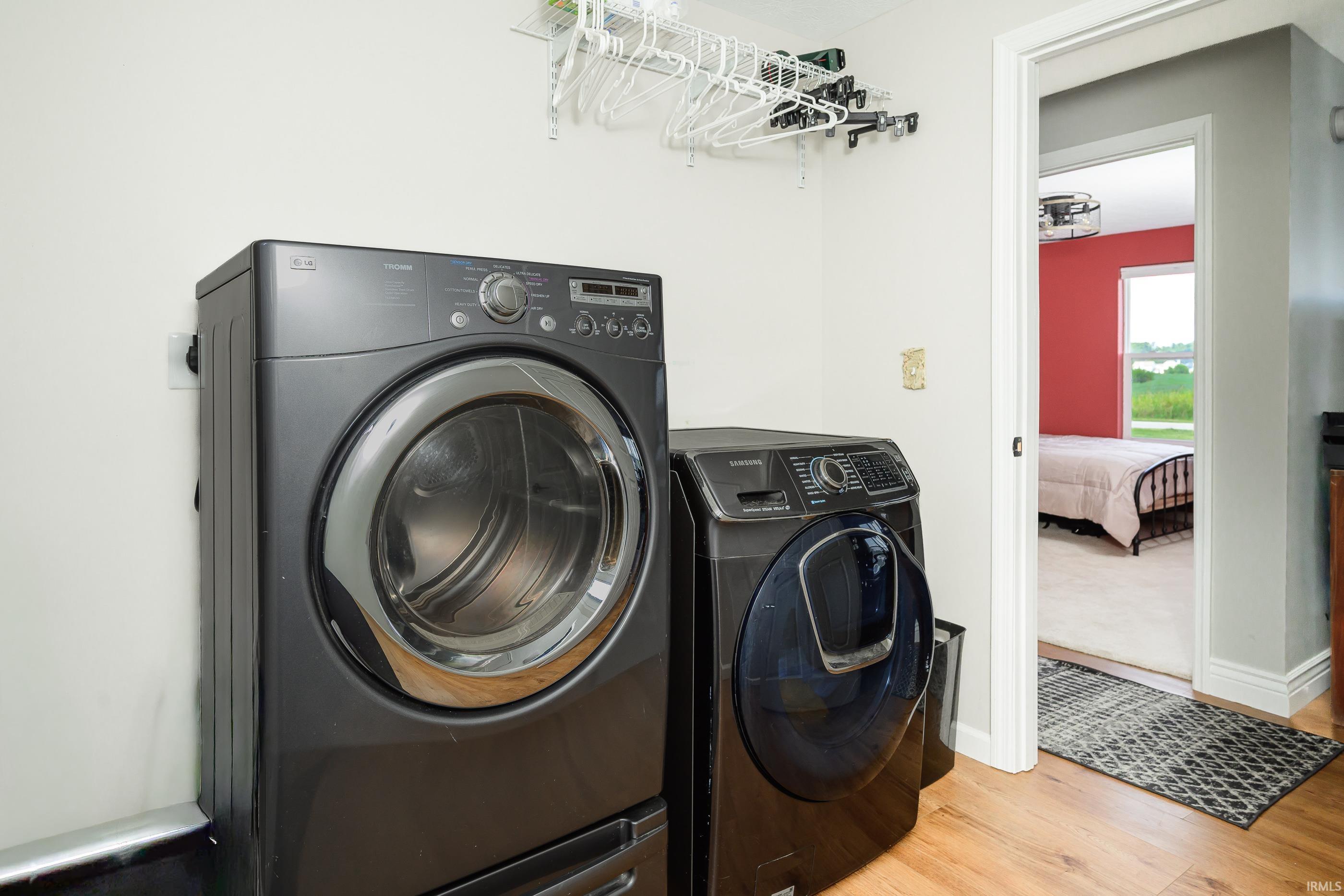 Laundry room with dark front-loading washer and dryer, wall-mounted clothes racks, and a bedroom visible through the doorway.