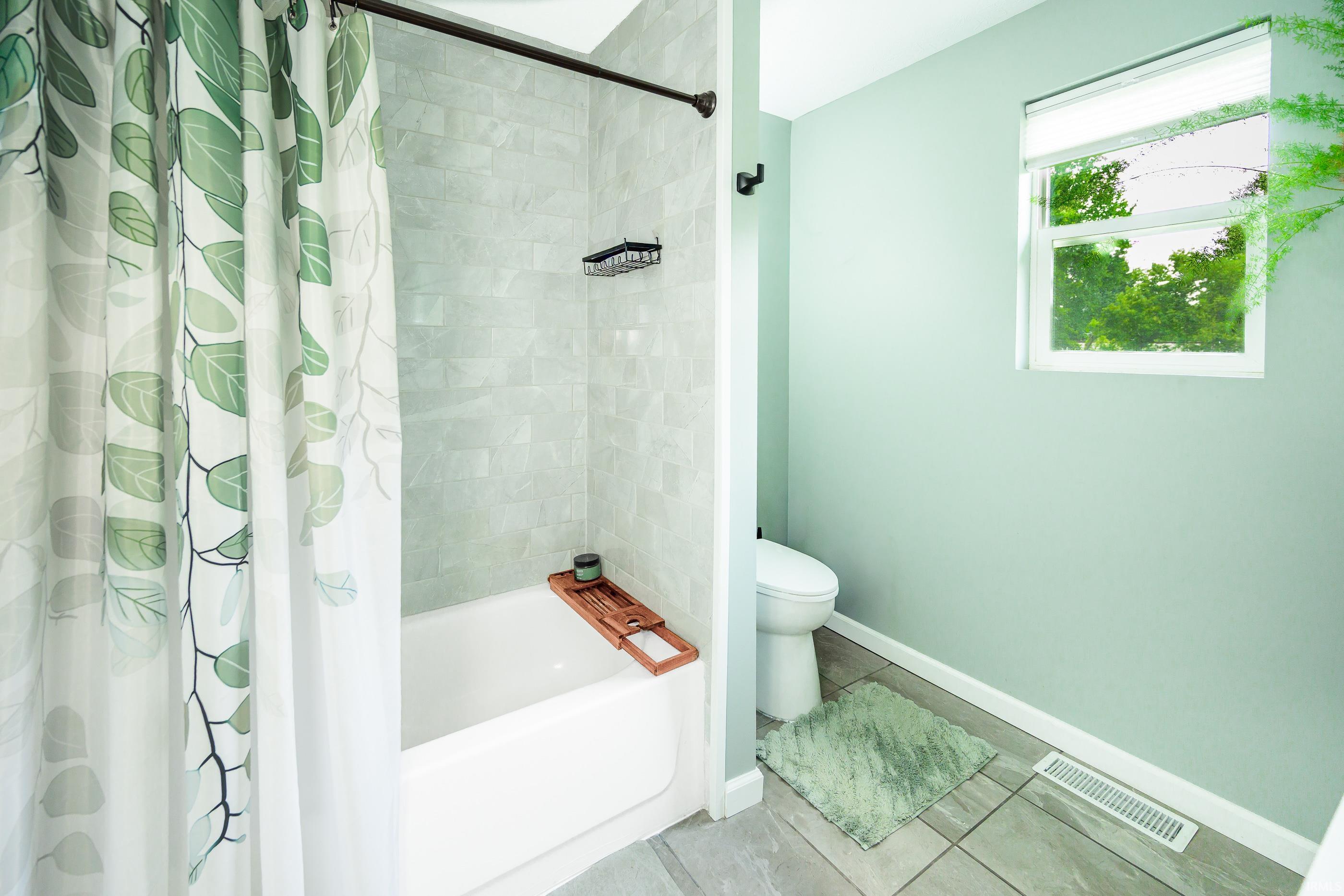 Bathroom with a tiled tub and shower, leafy curtain, and window overlooking trees.