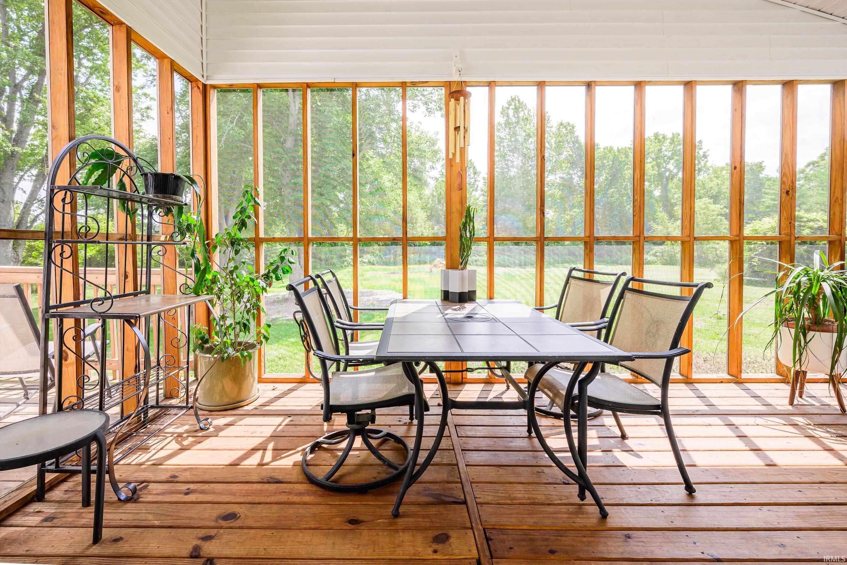 Furnished sunroom with tall wood-framed windows, potted plants, and a dining table overlooking a green yard.