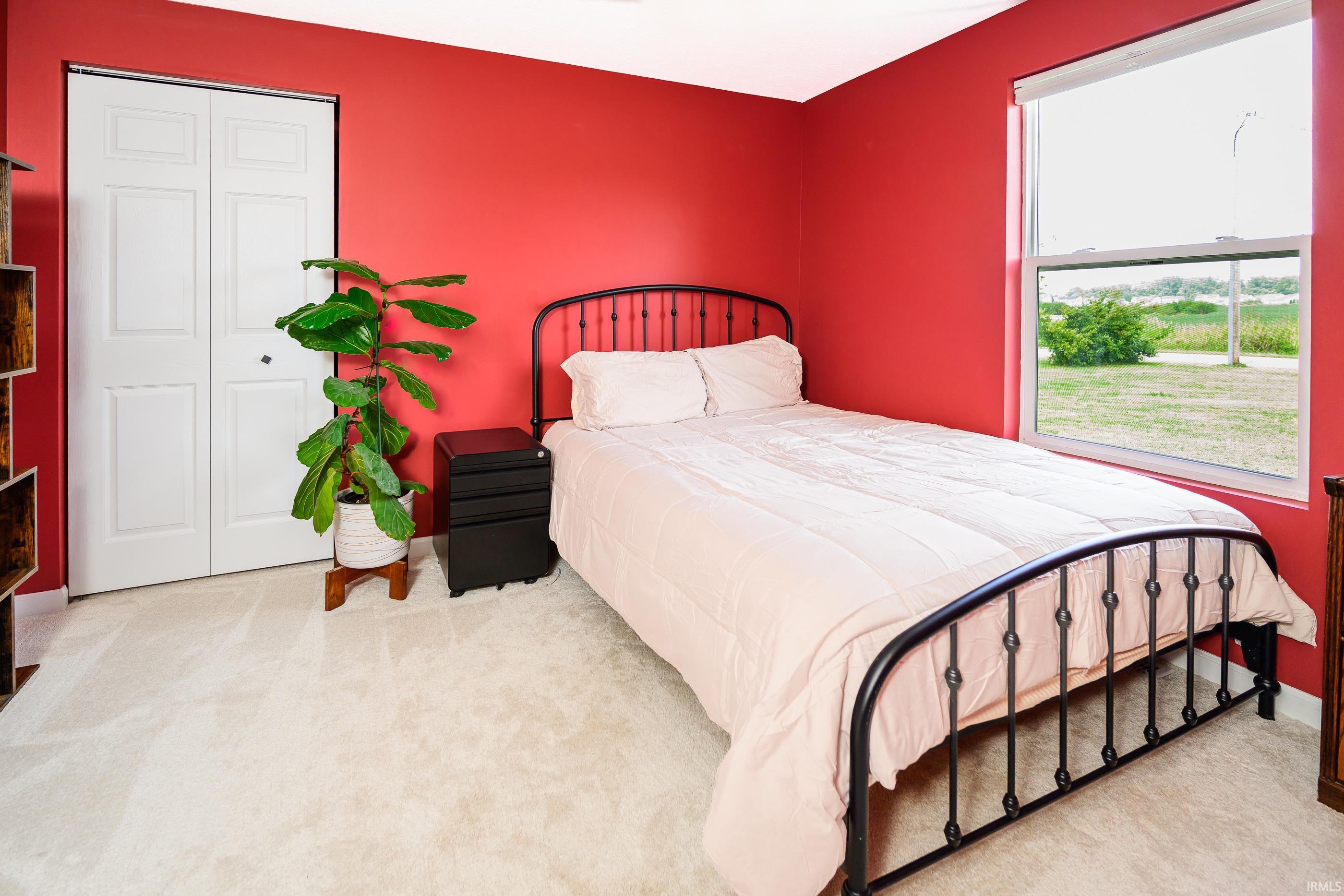 Bedroom with red walls, black metal bed, pale bedding, and a large window overlooking the yard.