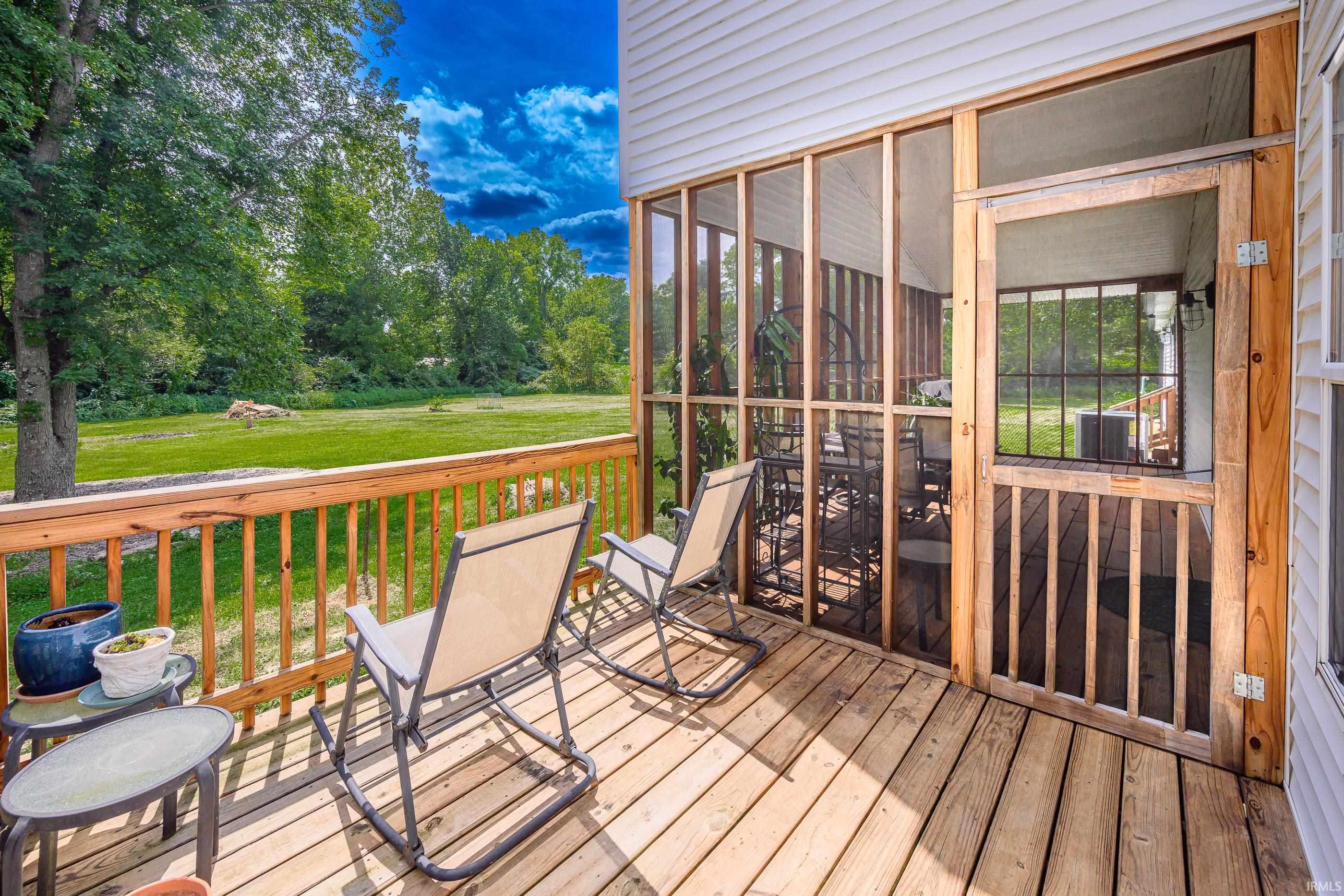 Wooden deck with rocking chairs beside a screened porch, overlooking a lawn and mature trees.