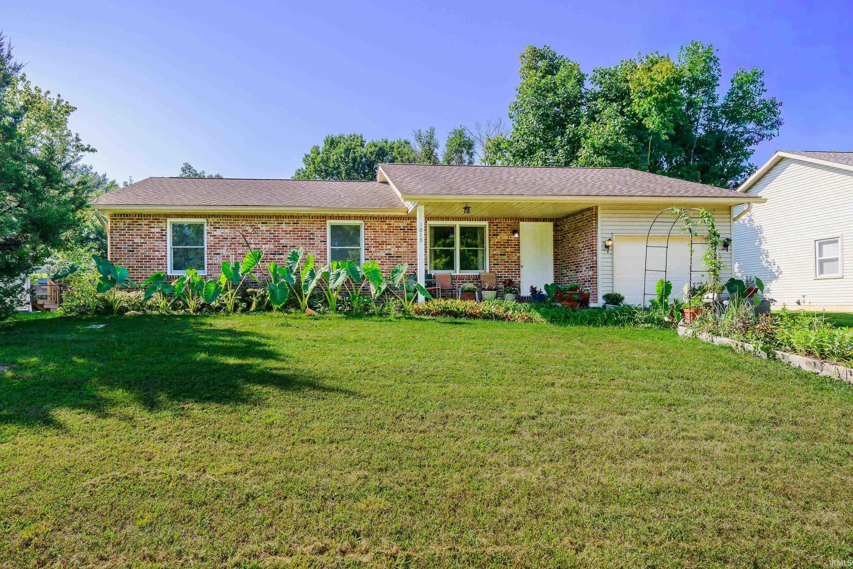 Single-story brick ranch with an attached garage, covered porch, and lush garden beds.