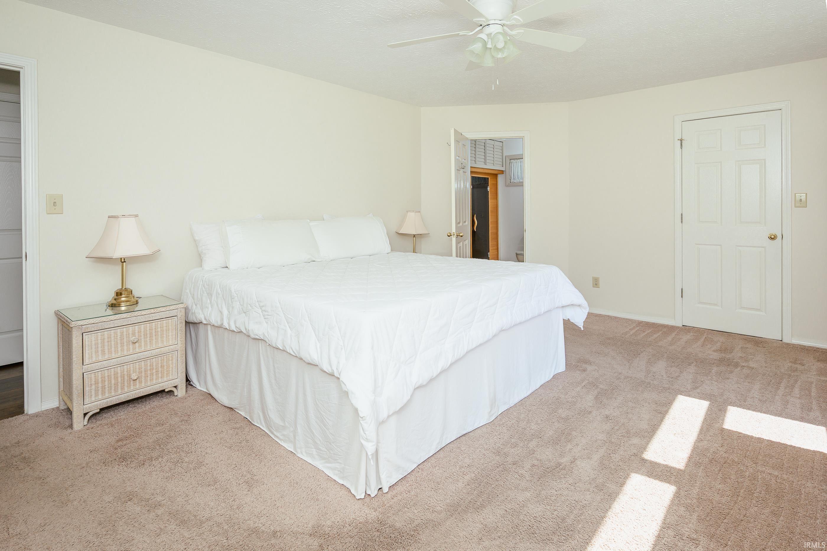 Bedroom with a large white bed, beige carpet, bedside lamps, and sunlight across the floor.