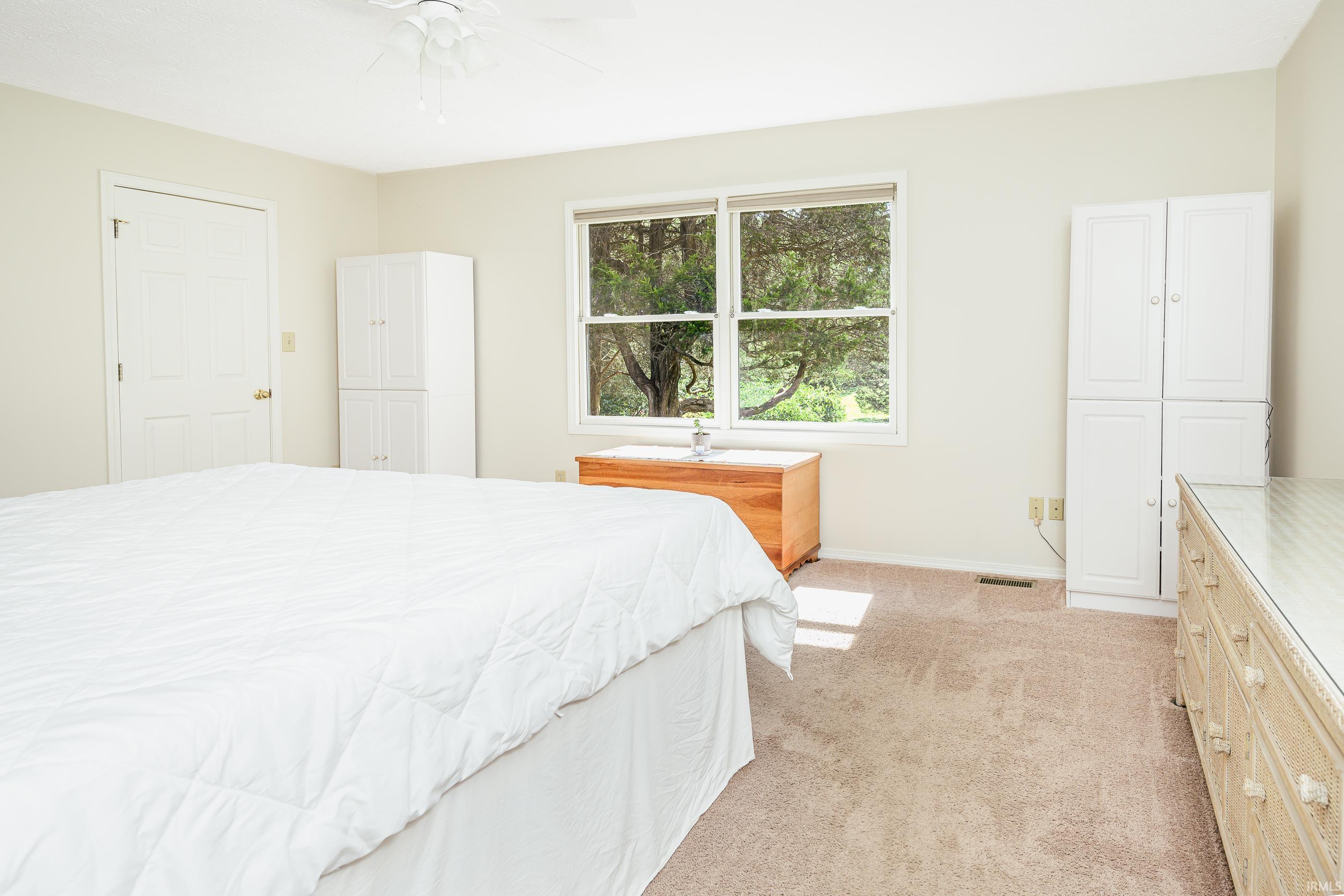 Bedroom with a white-covered bed, built-in cupboards, and a chest beneath a window overlooking trees.