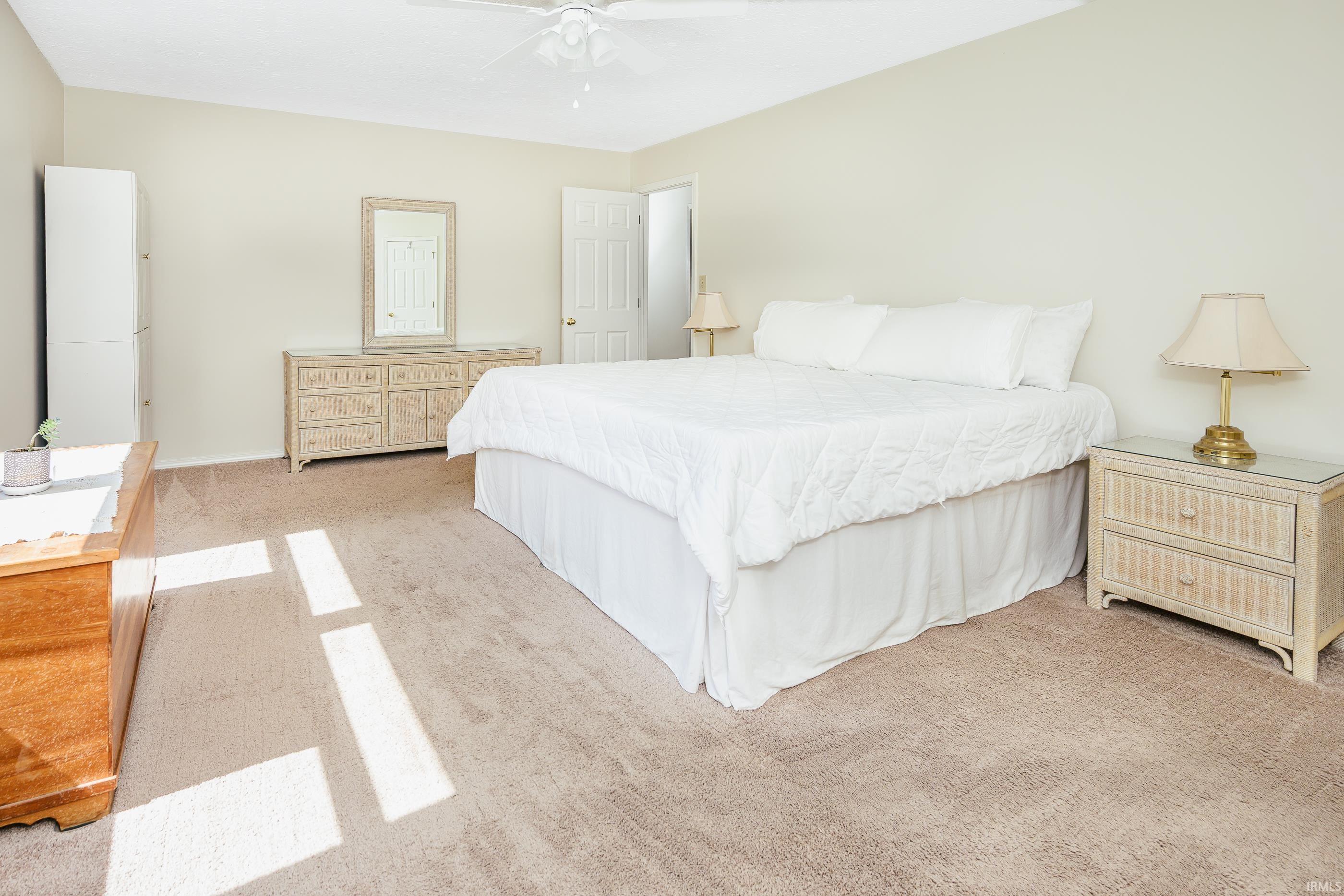 Bright bedroom with a large white bed, wicker-style furniture, and sunlight across the carpet.