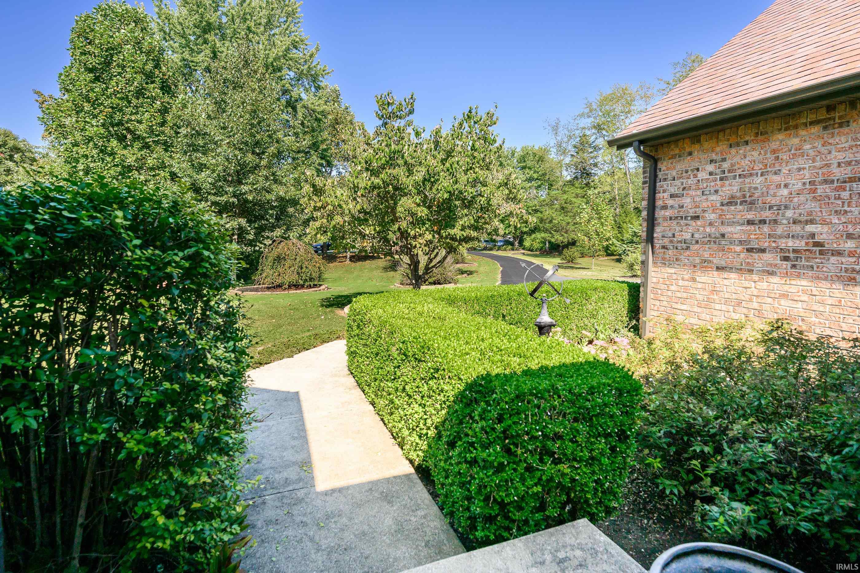 Concrete walkway bordered by trimmed hedges beside a brick home and leafy trees.