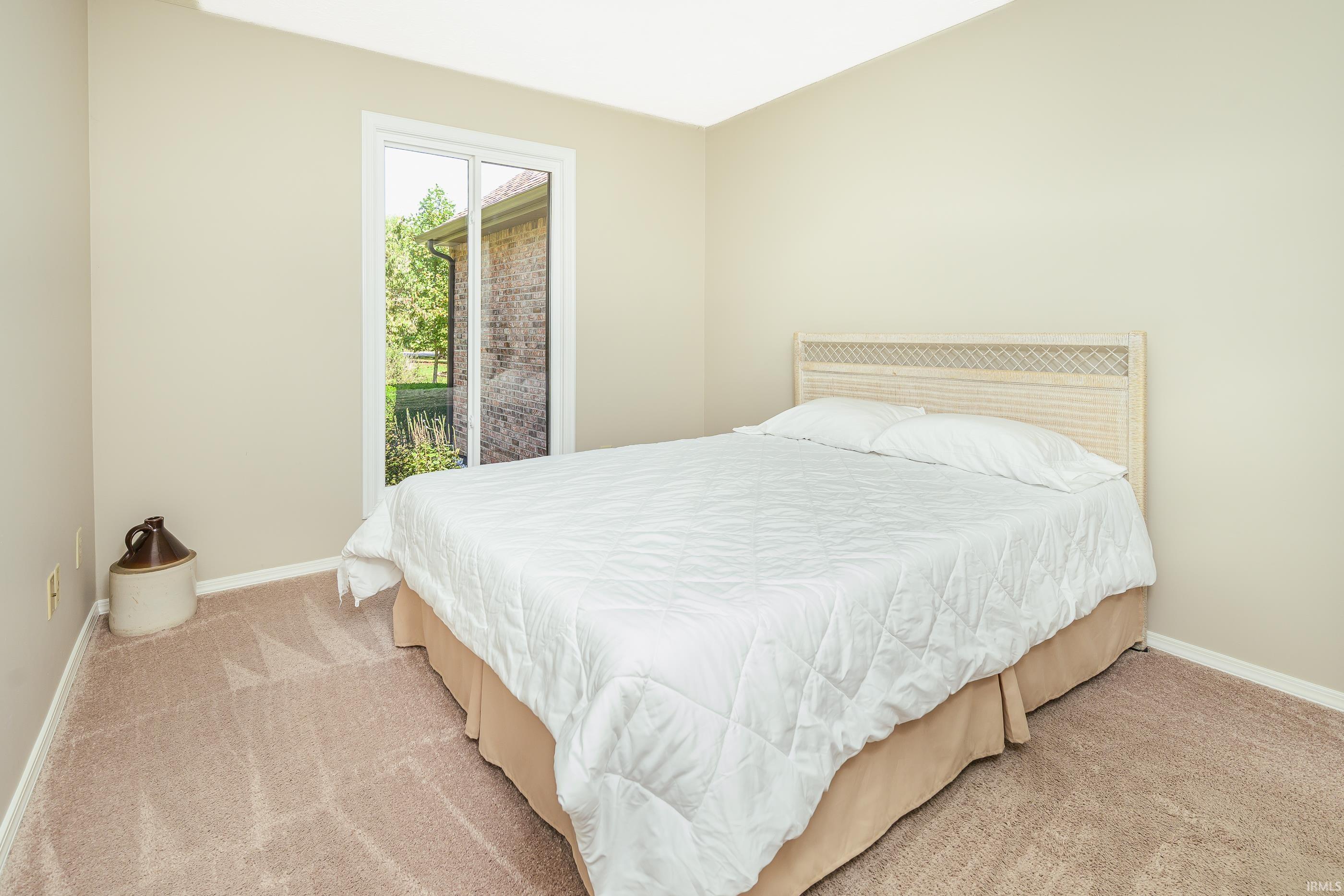 Staged bedroom with a white-covered bed, woven headboard, beige carpet, and tall window.