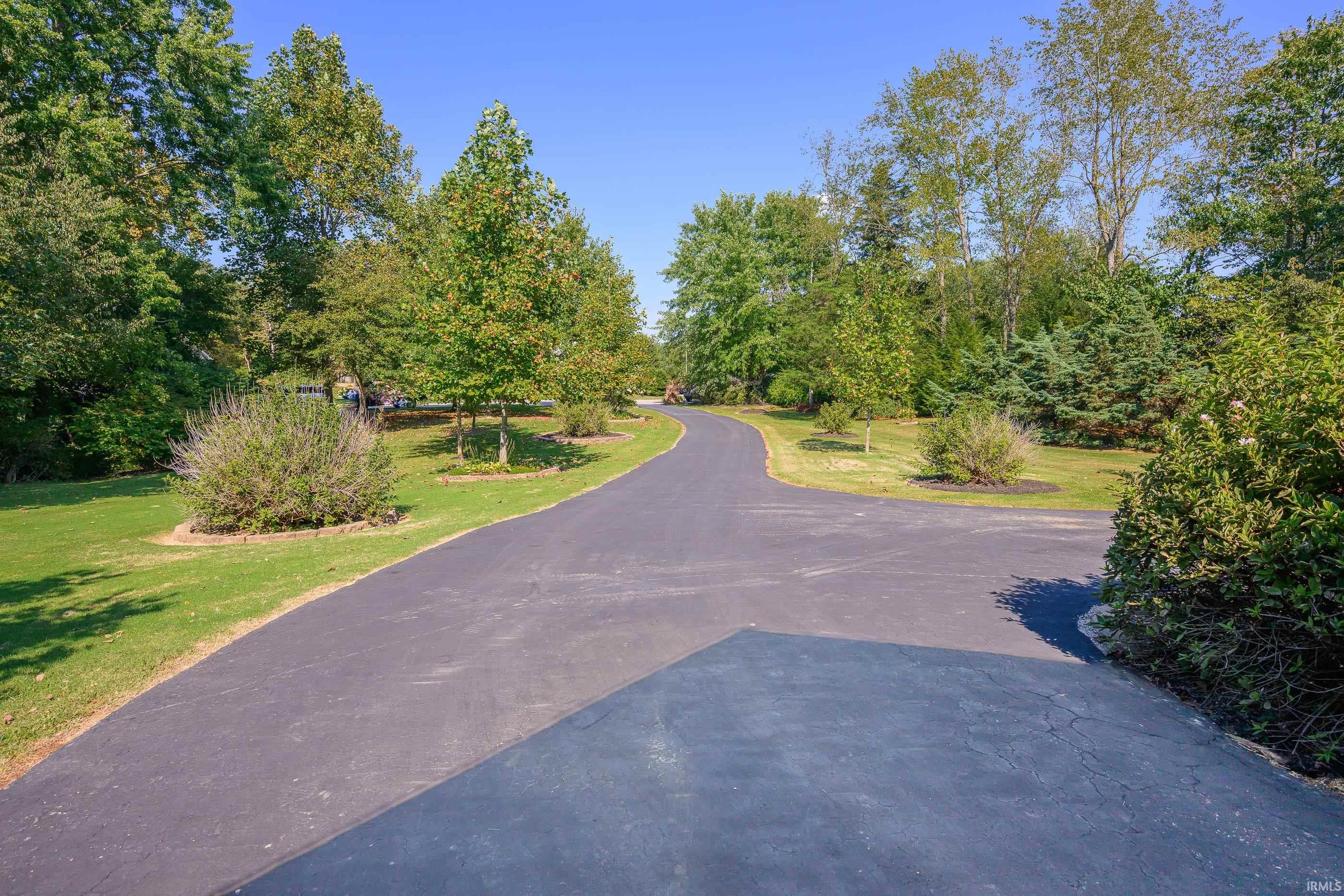 Curving paved driveway bordered by lawns, mature trees, and landscaped shrubs.