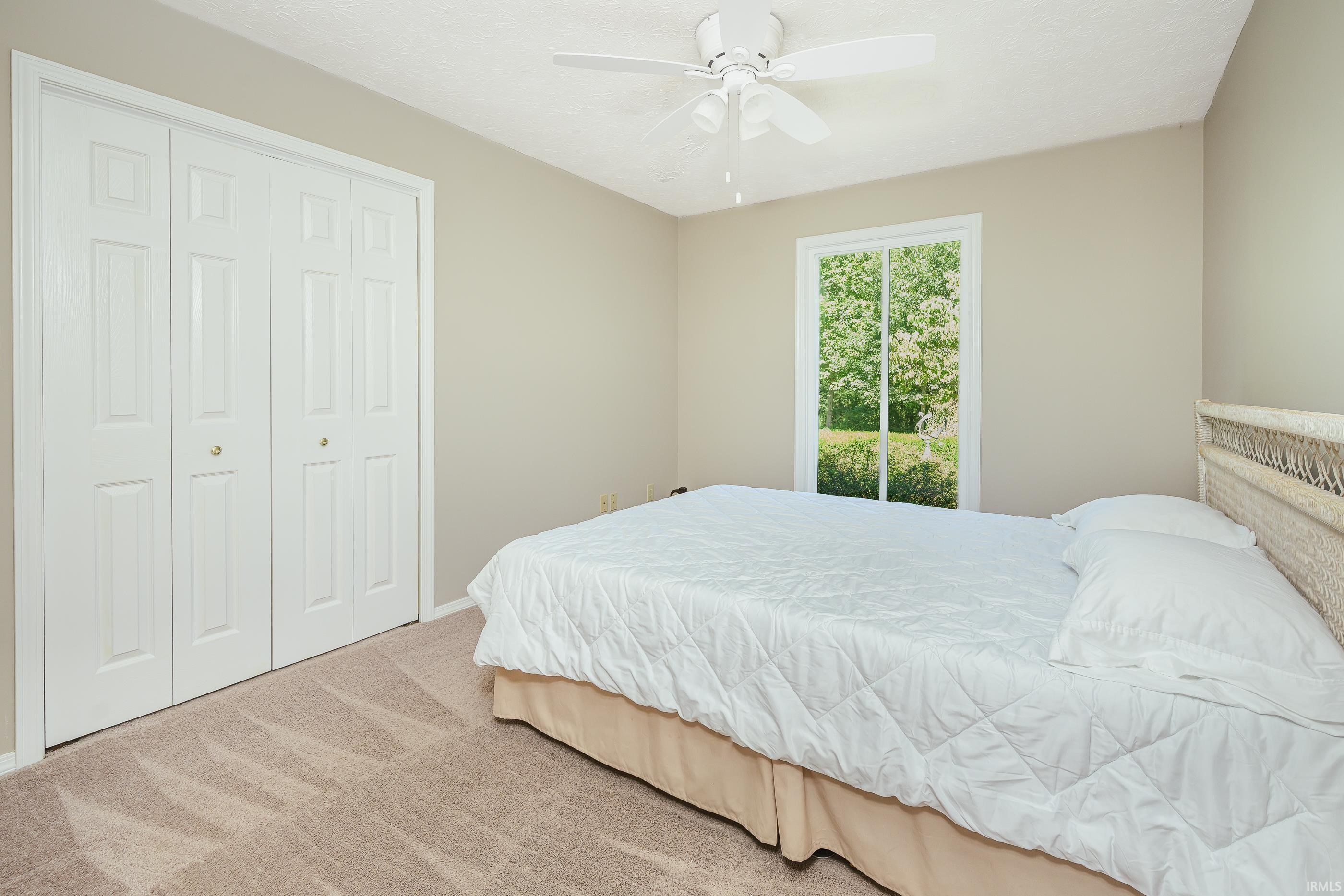 Bedroom with a white quilted bed, ceiling fan, and window overlooking greenery.