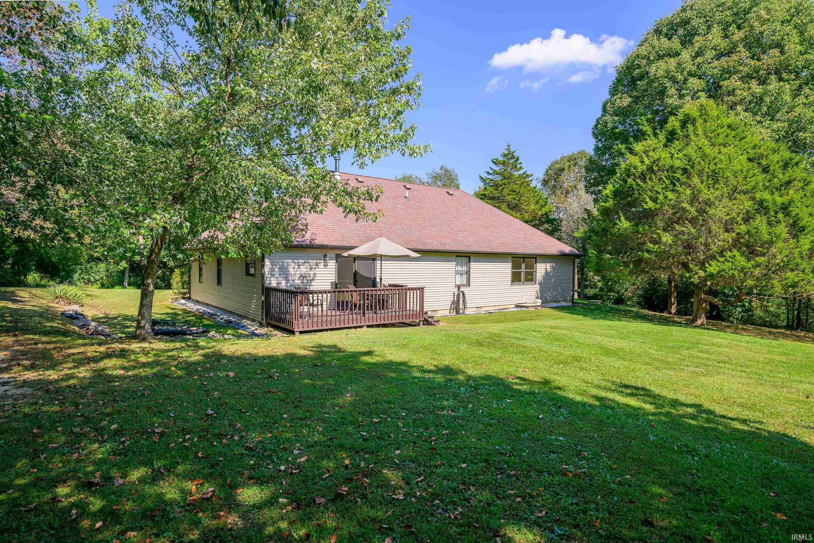 Rear of a single-story home with a small deck, broad lawn, and mature trees.