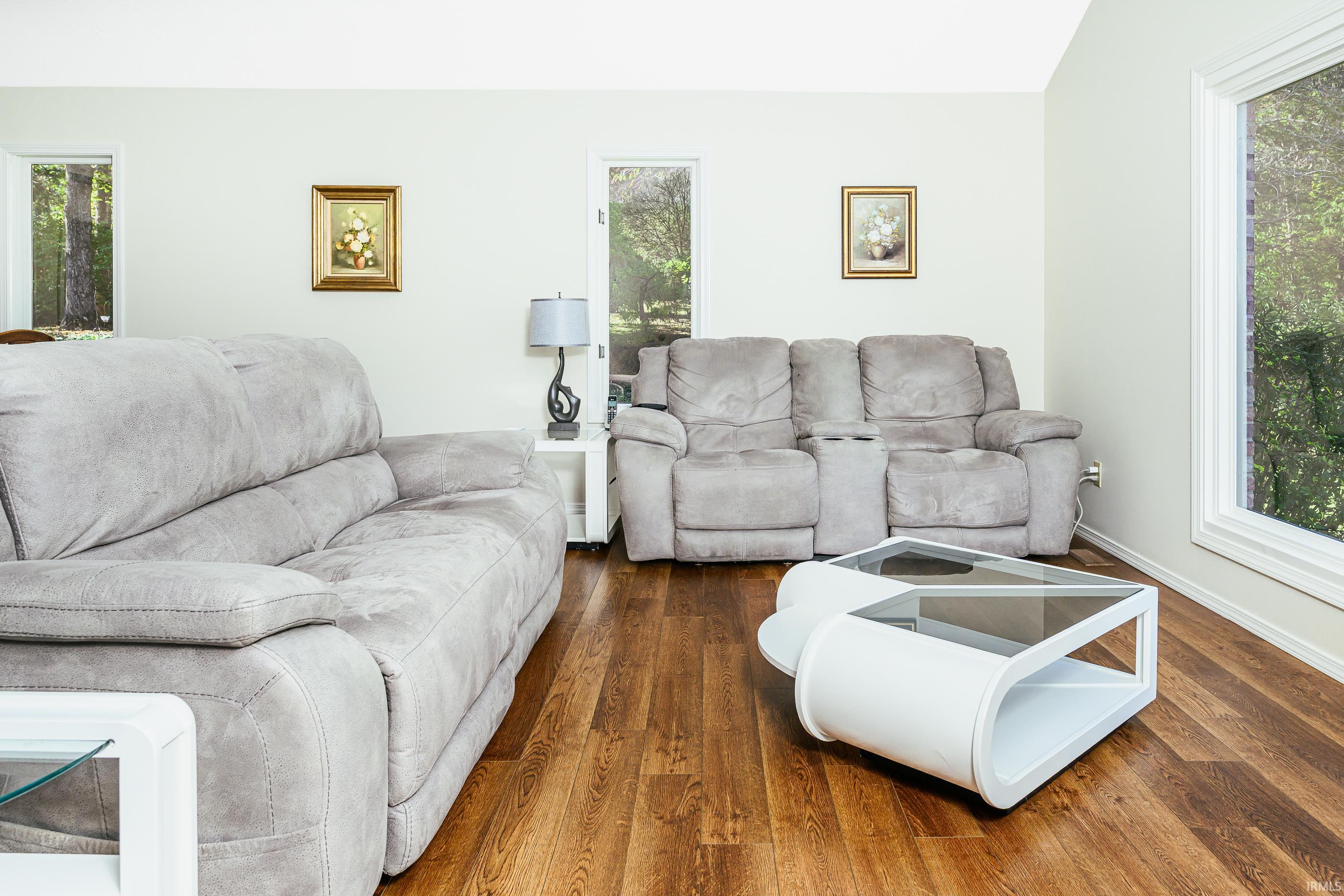 Living room with gray reclining sofas, hardwood floors, and large windows overlooking trees.