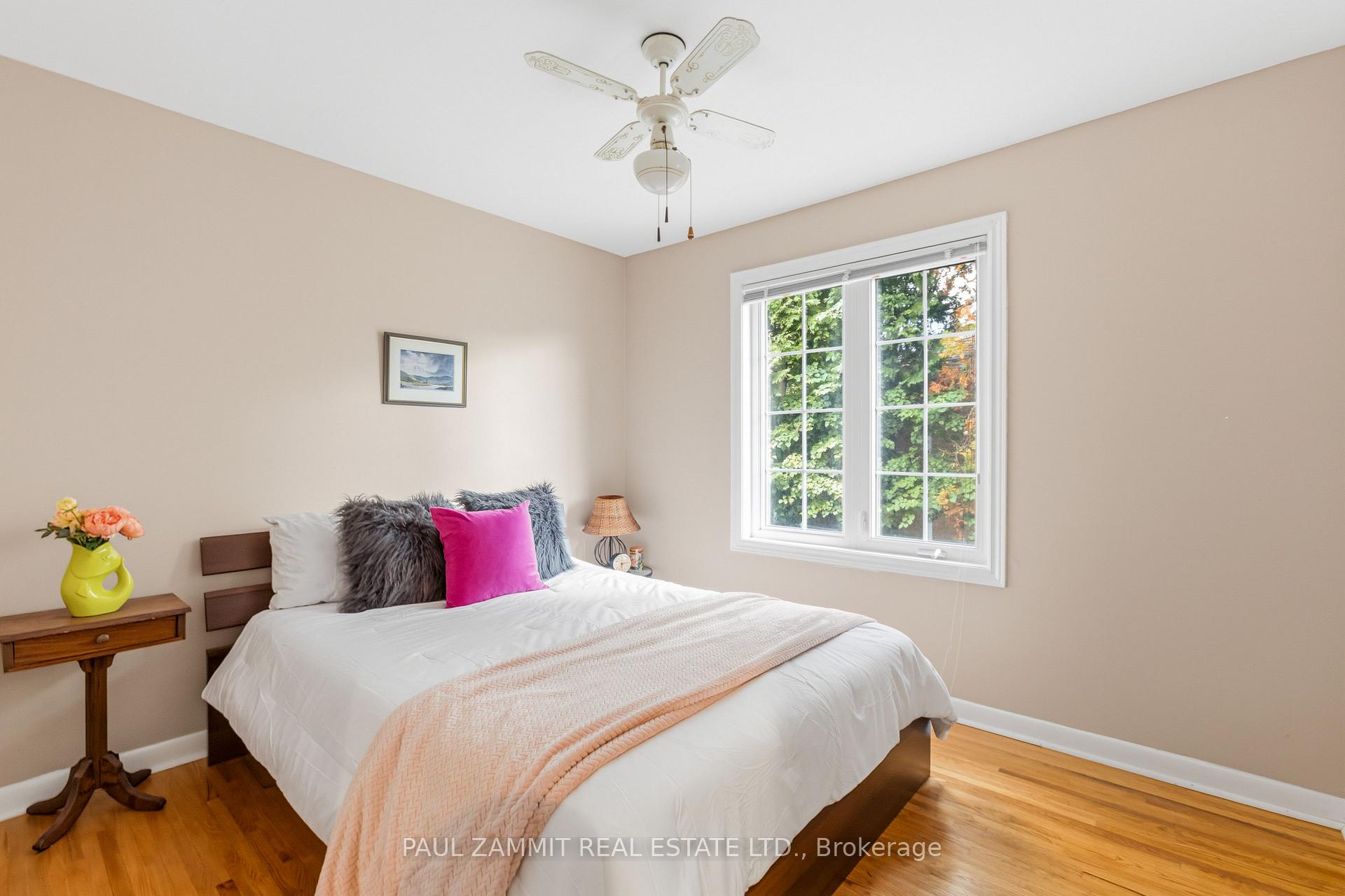Staged bedroom with a white bed, bright pink accent pillow, and large window overlooking leafy trees.
