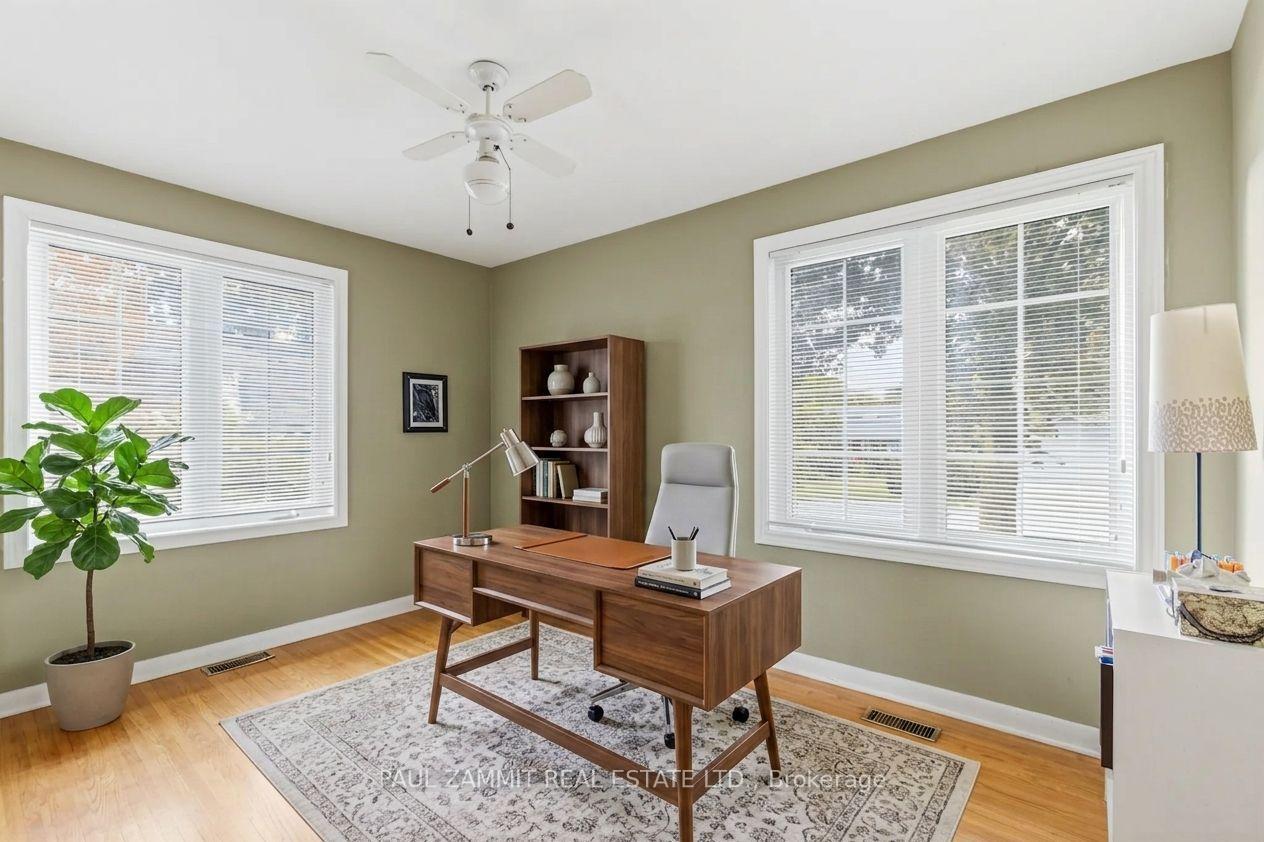 Furnished home office with a wooden desk between two large windows with blinds.
