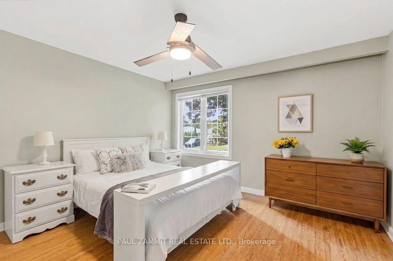 Staged bedroom with a white bed, wooden dresser, hardwood floors, and a window overlooking greenery.