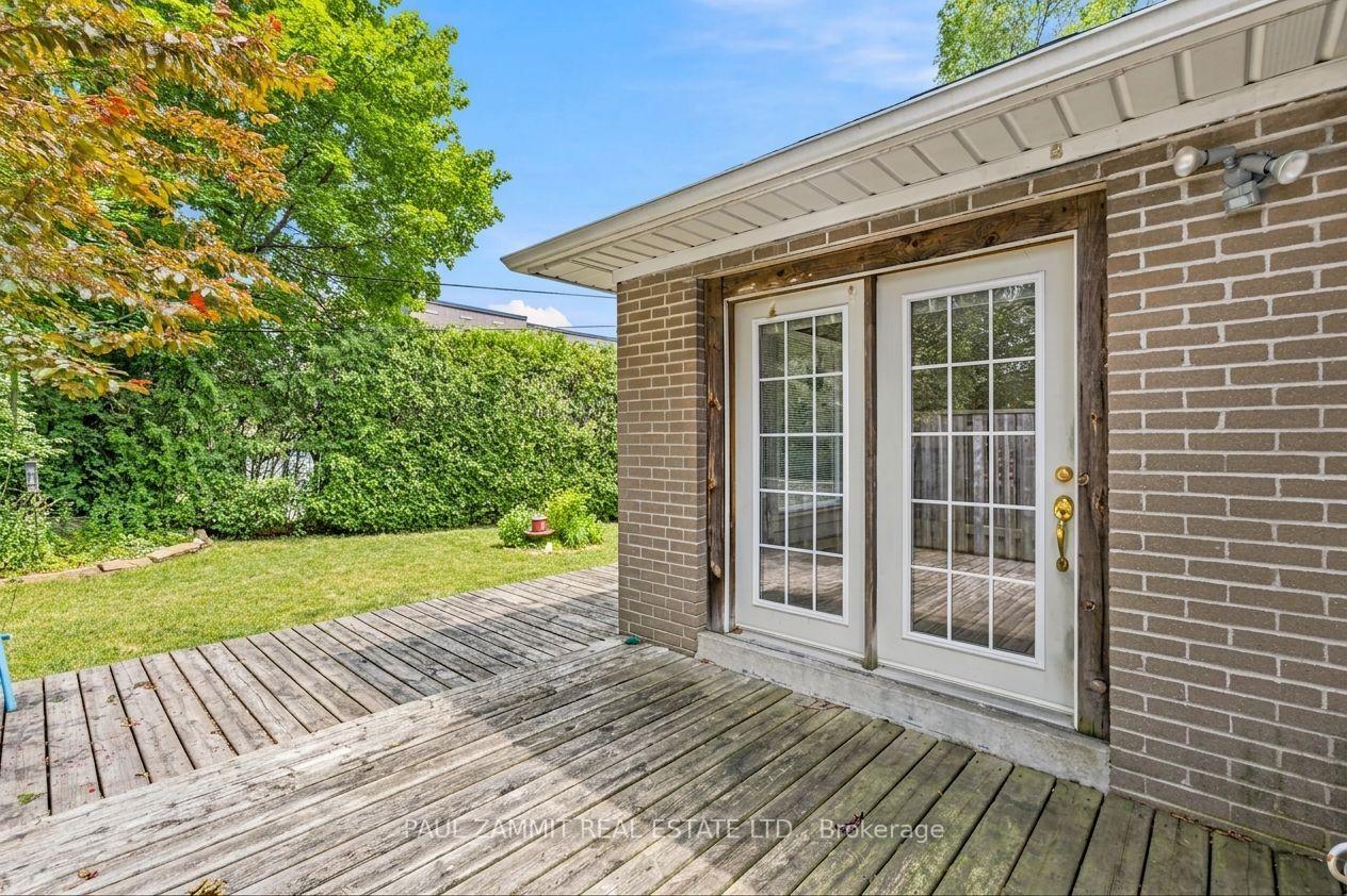 Backyard deck beside a brick house, with white patio doors, lawn, hedges, and mature trees.