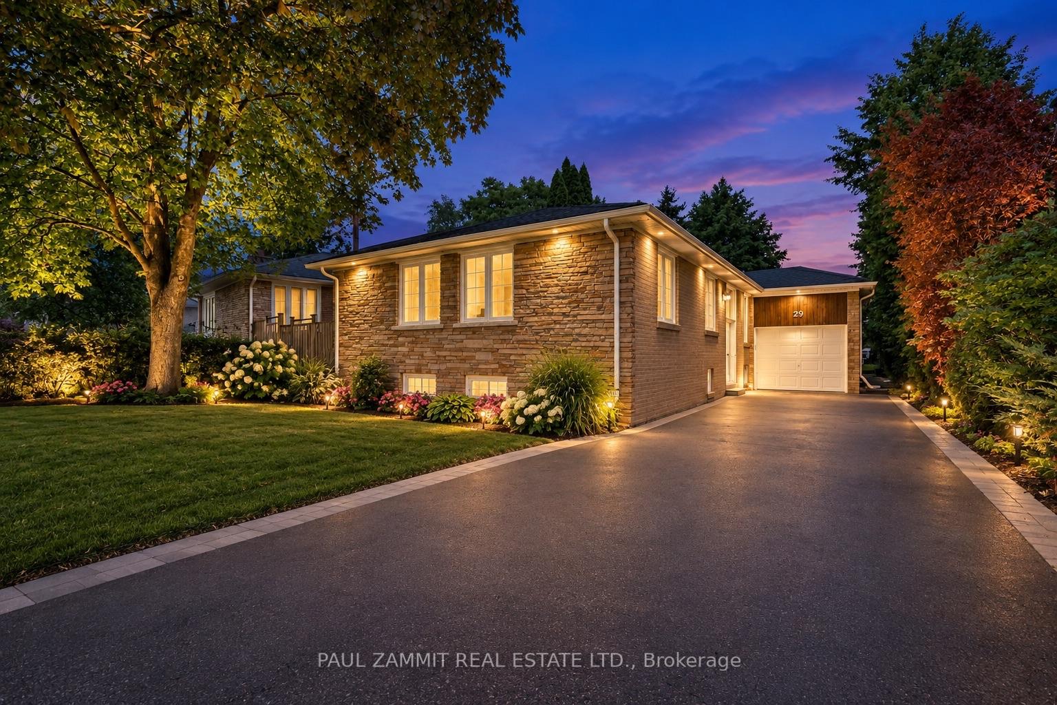 Stone-clad house at dusk with glowing windows, landscaped flower beds, and an attached garage.