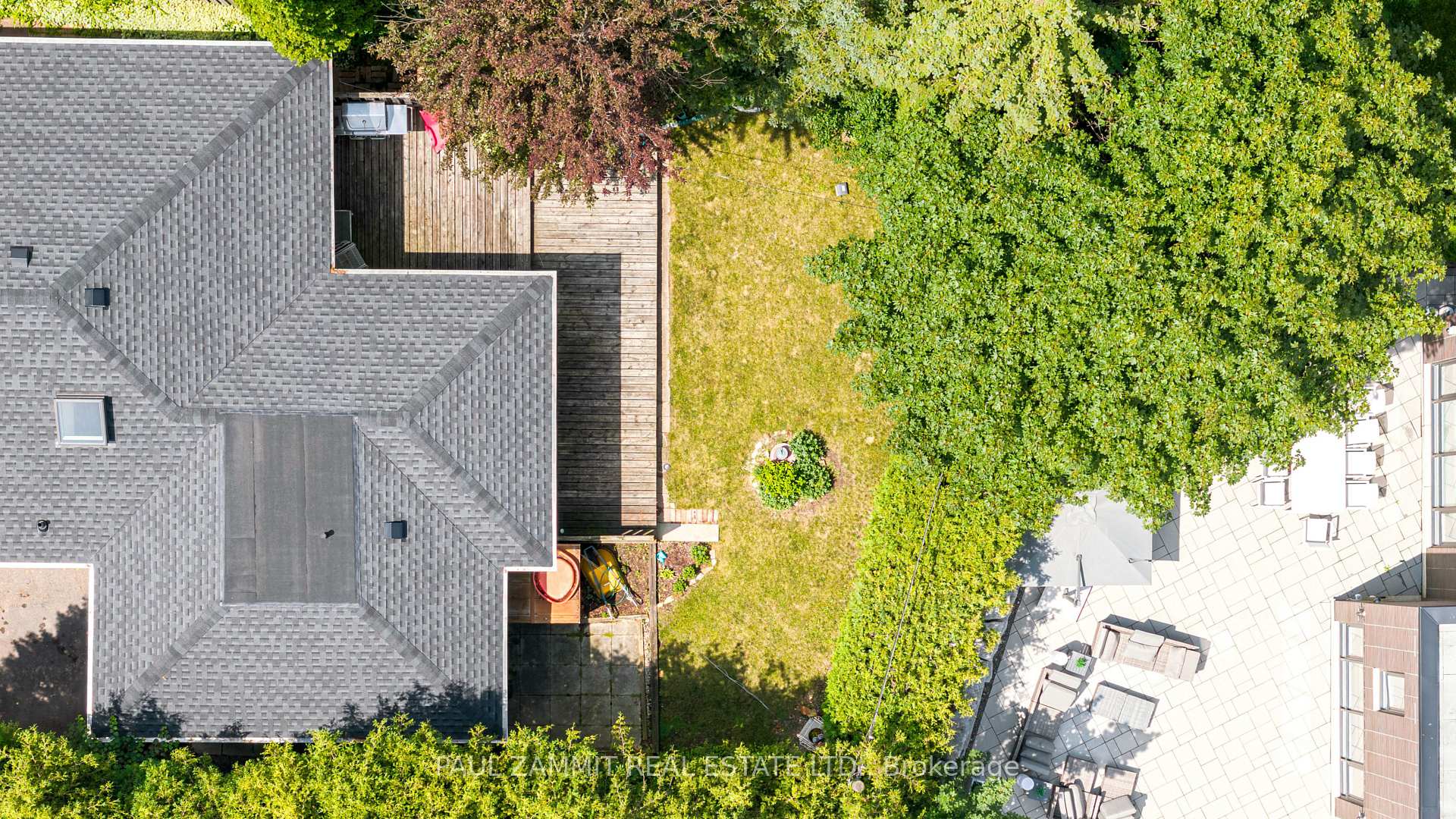 Aerial view of a home and backyard with a wooden deck, lawn, mature trees, and paved patio.