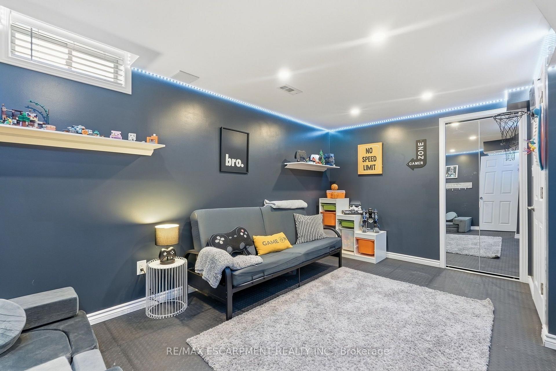 Furnished gaming room with blue walls, LED lighting, game-themed cushions, collectibles, and a basketball hoop.