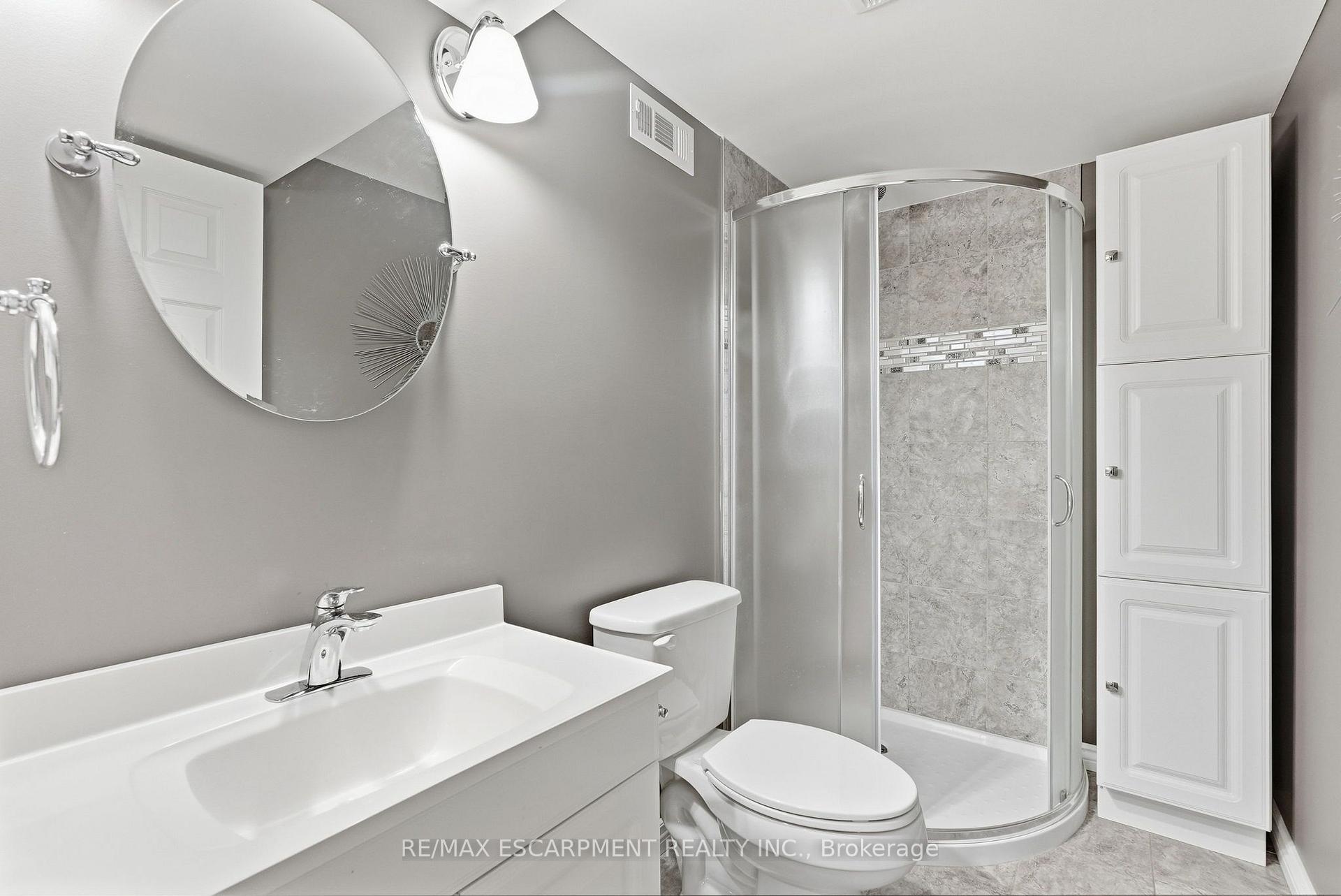 Bathroom with a curved glass shower, gray tile, white vanity, and tall storage cabinet.