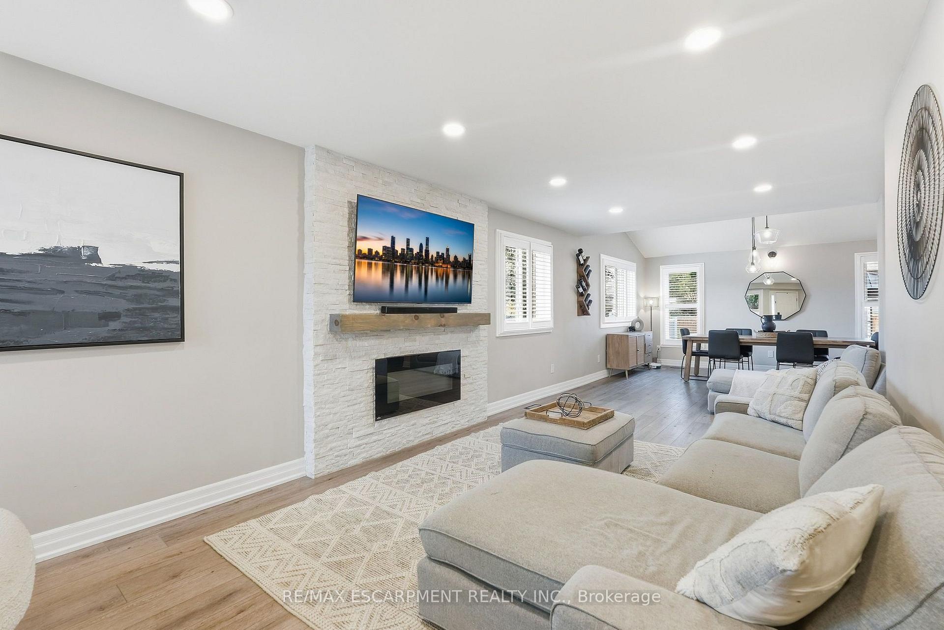 Open-plan living and dining room with a white stone fireplace, mounted TV, neutral sectional, and light wood floors.