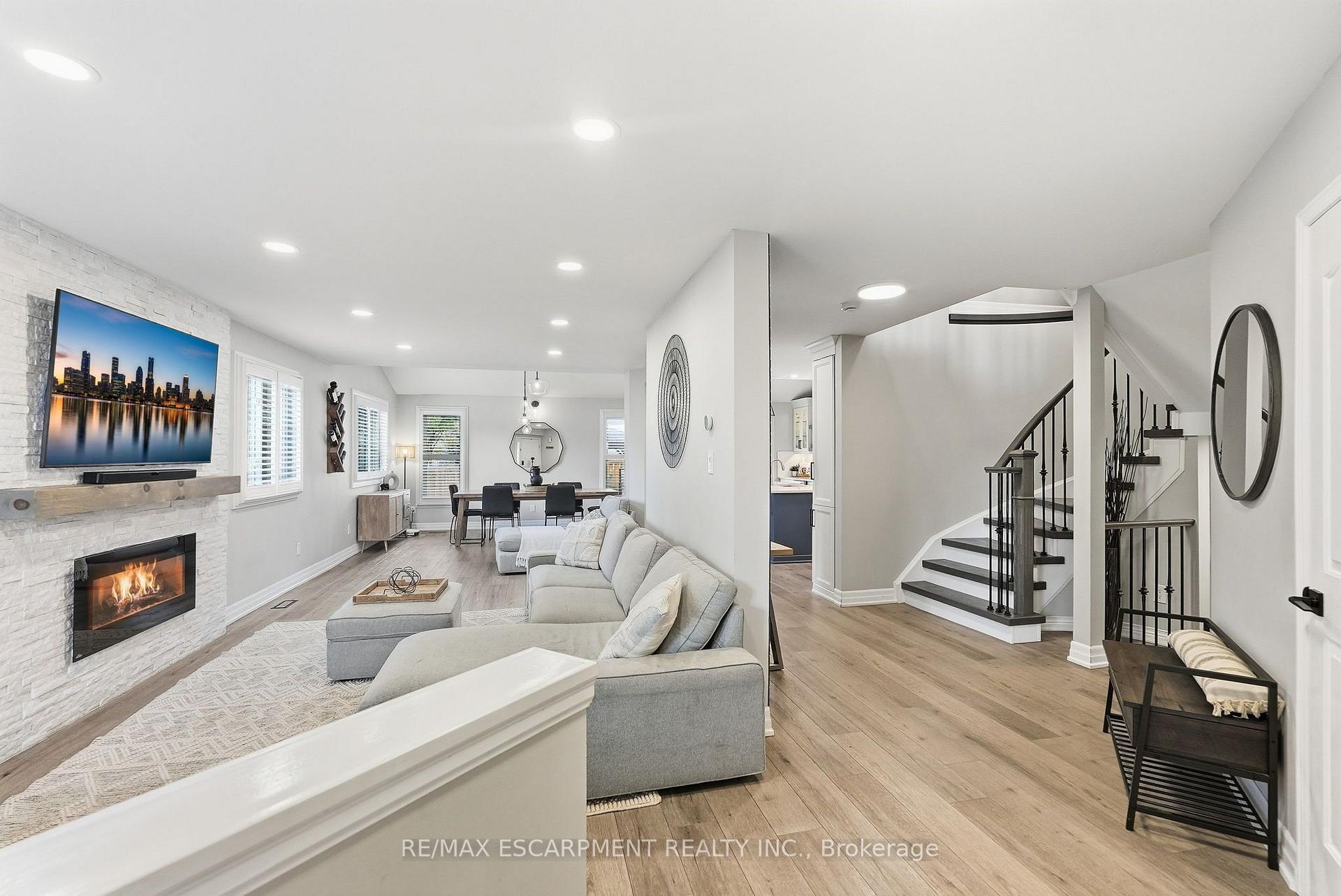 Open-concept living room with a stone fireplace, gray sectional, dining area, and staircase.