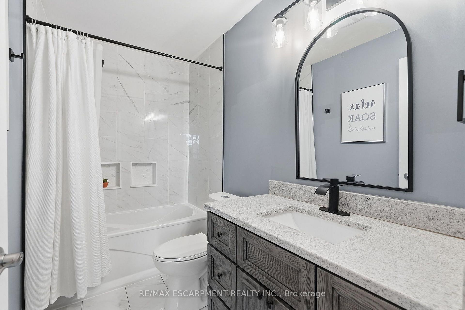 Bathroom with marble-look tub surround, gray wood vanity, black faucet, and arched mirror.